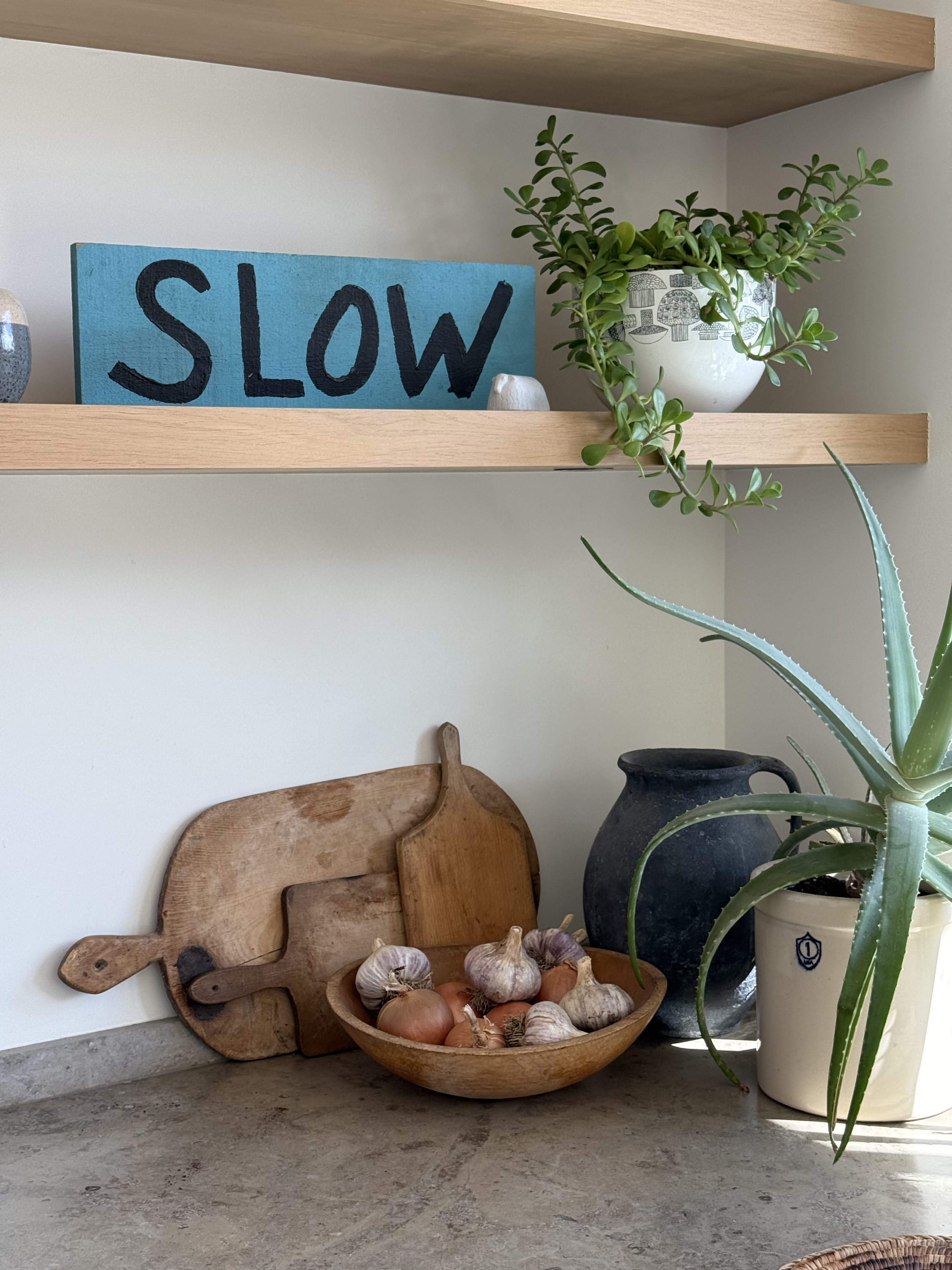 Wooden shelves with a potted succulent, a black vase, a vase with a blue pattern, a light blue sign reading 'SLOW', dried garlic and onions in a bowl, and wooden cutting boards, all against a pale wall and a concrete countertop.