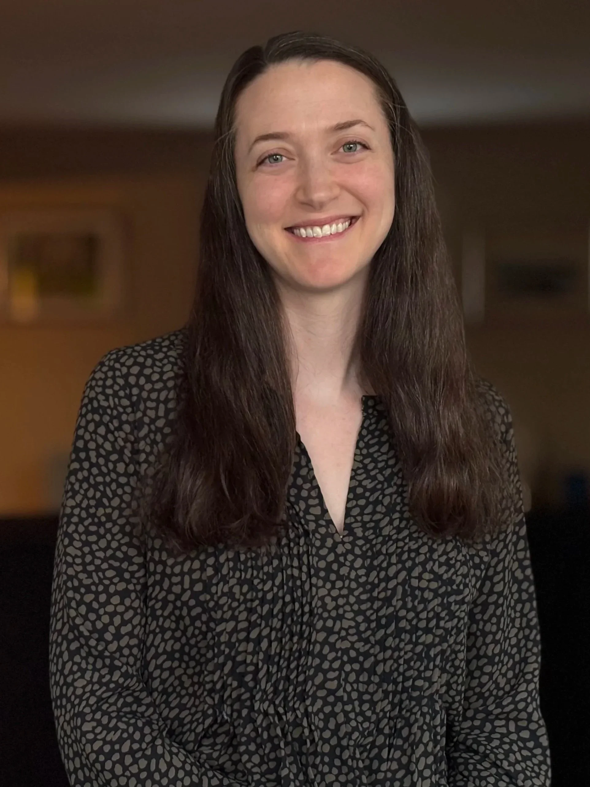 A woman with long brown hair, wearing a black and white patterned blouse, smiling at the camera in a warmly lit room.