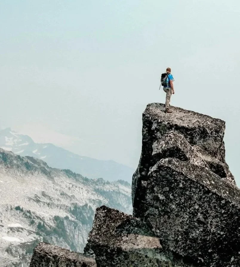 A person with a backpack standing on top of a large rock formation at the edge of a mountain, with a mountain range in the background.