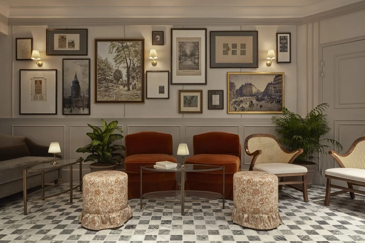 A cozy sitting room with a wall decorated with framed artwork, including paintings and photographs. There are several chairs, including two plush orange armchairs, one cane-back chair, and a sofa, arranged around small tables with lamps. Potted plants are on either side of the seating area, and the floor is tiled with a patterned black and white design.