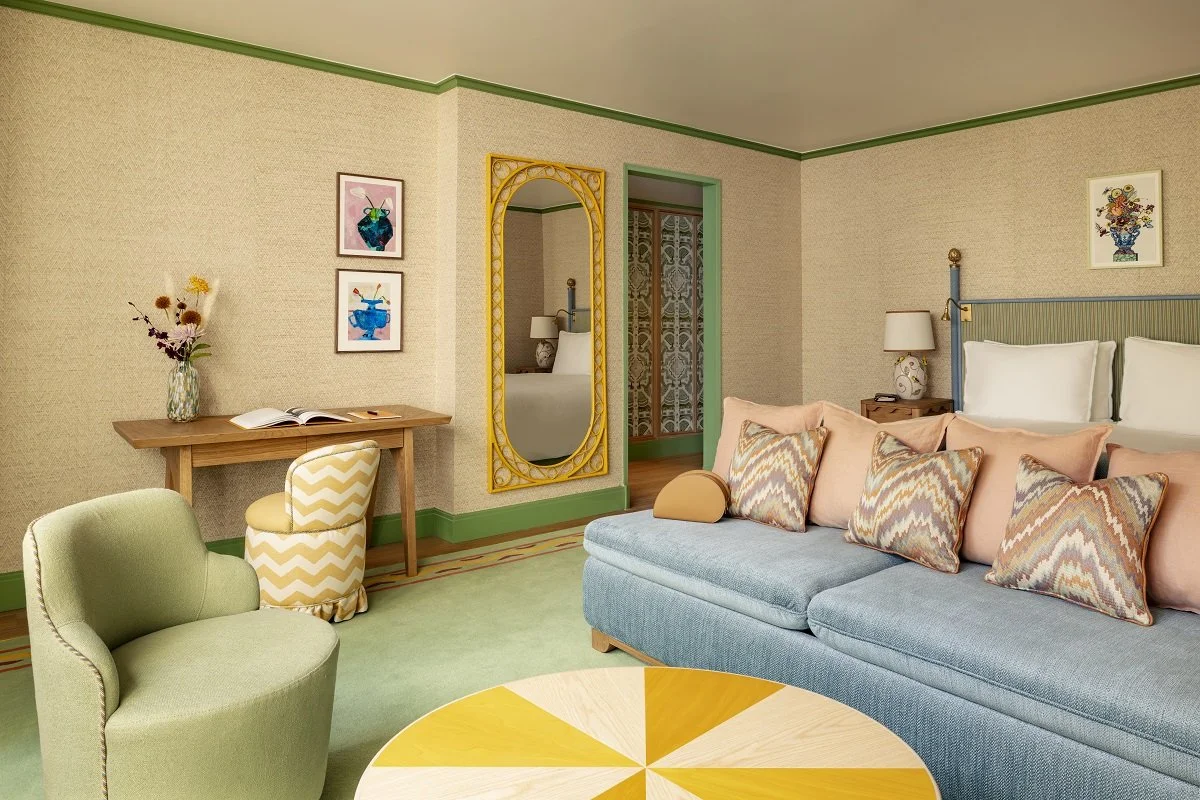 A cozy hotel room or living space with a light green armchair, a multi-colored patterned sofa with pillows, a wooden desk with a vase of flowers, a mirror, a bedside table with a lamp, and colorful artwork on beige walls with green trim.