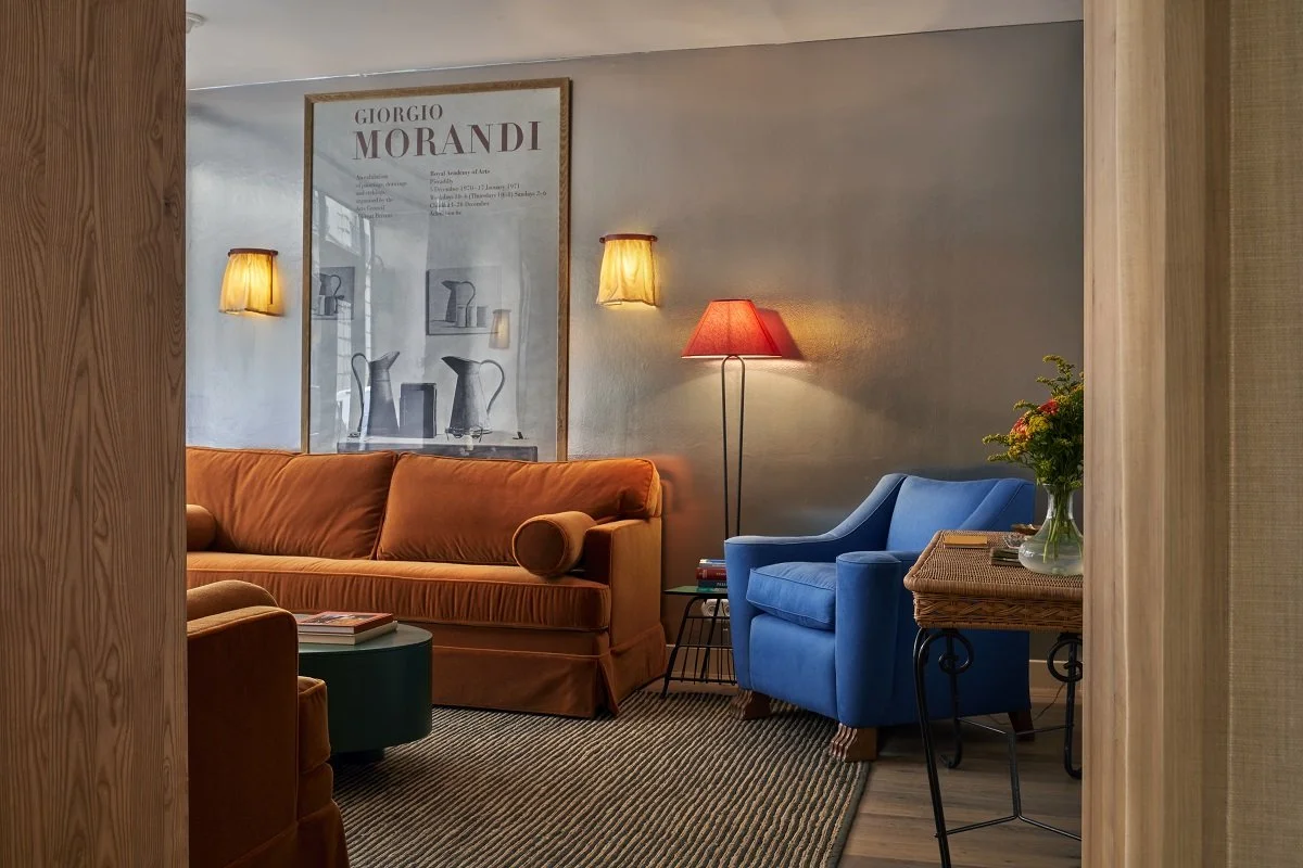 A cozy living room with a burnt orange sofa, a blue armchair, a small side table with a vase of flowers, and wall-mounted lamps.
