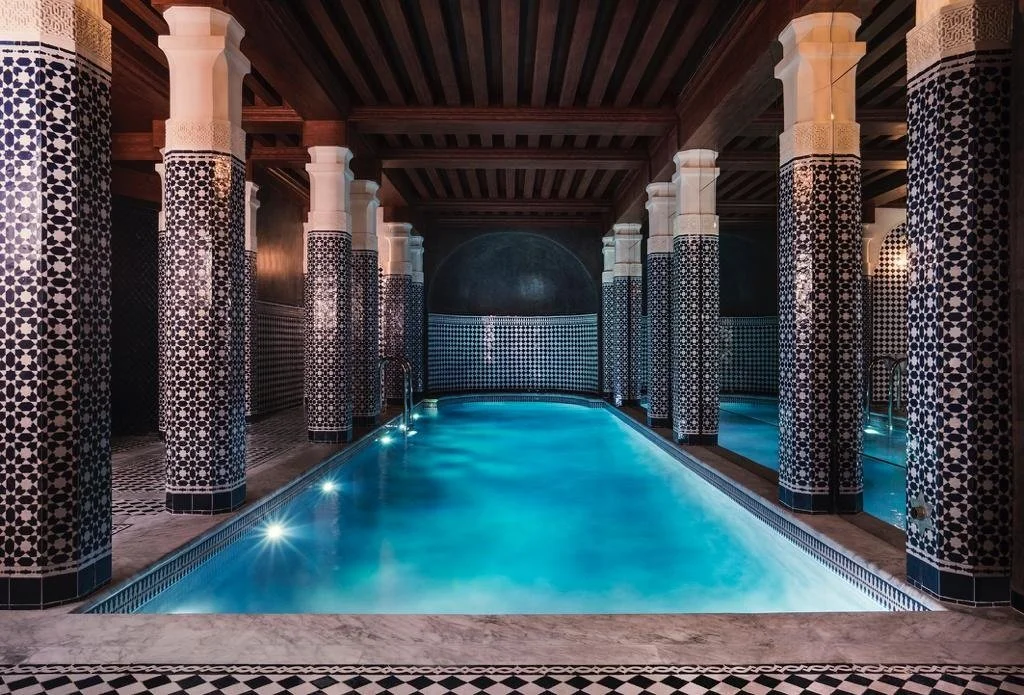 Indoor swimming pool with ornate Moroccan-style black-and-white tile columns and walls, illuminated by underwater lights, with a wooden ceiling.