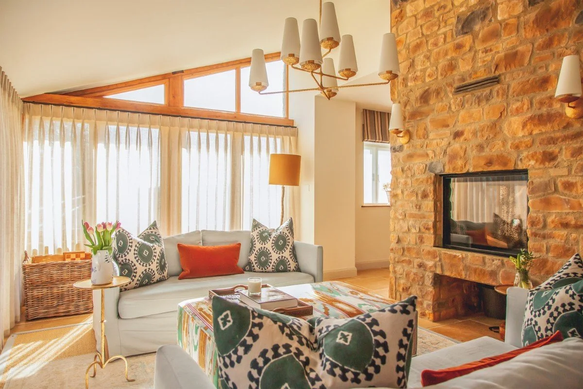 Living room with a stone fireplace, white sofas, patterned and orange pillows, a colorful coffee table, and large windows with curtains and a wooden valance.