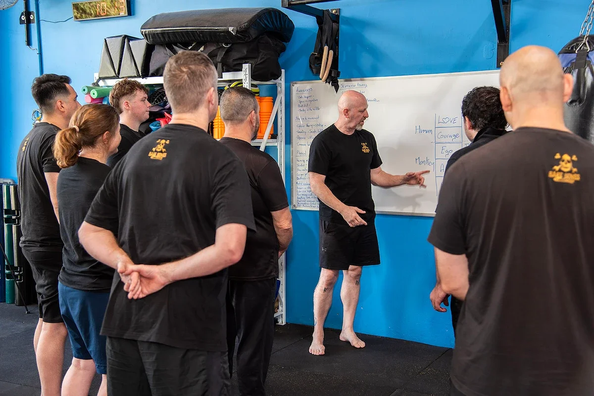 Jim Armstrong teaching BareBones Combatives concepts at a whiteboard during a self-protection workshop.