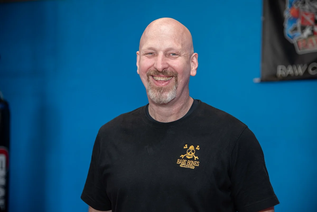 Jim Armstrong, founder of BareBones Combatives, smiling in a BareBones Combatives instructor shirt.