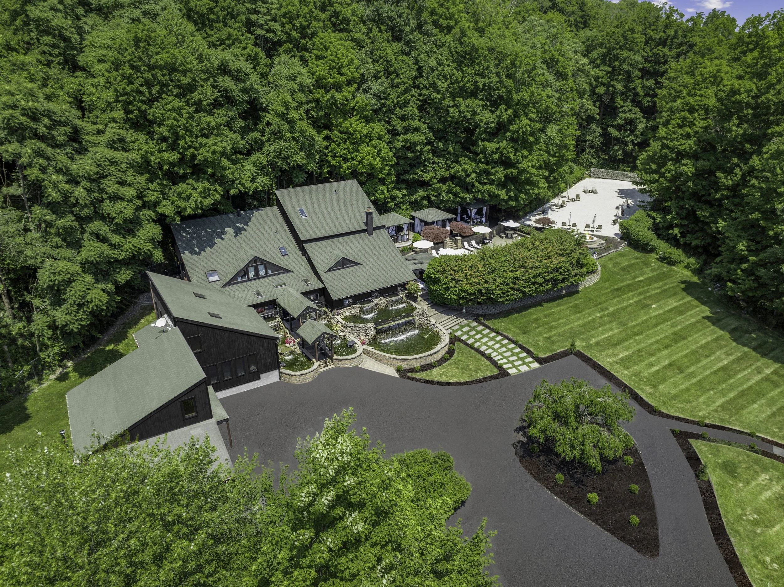 Aerial view of a house surrounded by lush green trees, with a driveway, multiple outdoor seating areas, and a landscaped garden.