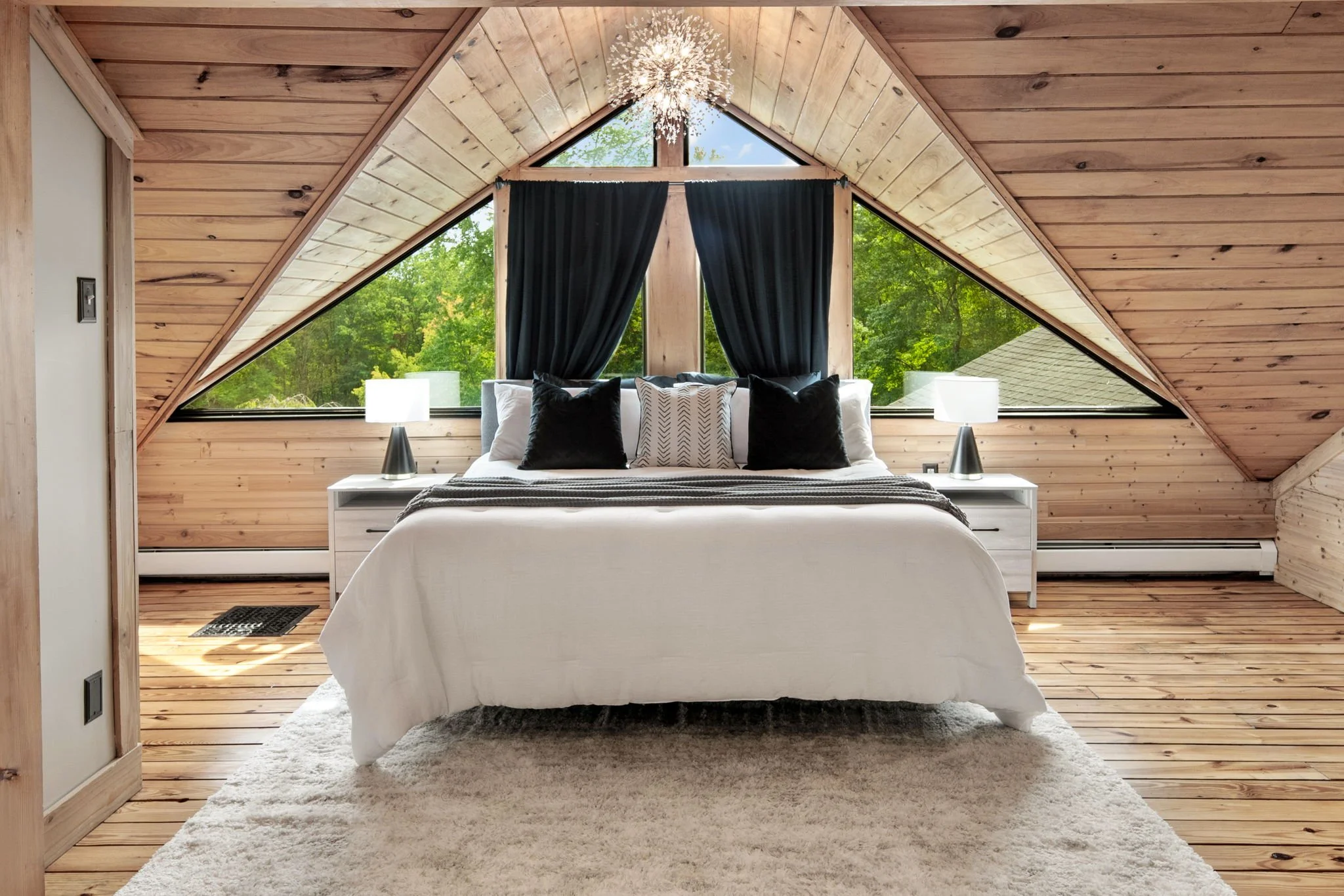 A cozy bedroom with a wooden ceiling and large triangular windows showing green trees outside. The bed has white bedding and black and white pillows. Two bedside lamps are on white nightstands, and a fluffy white rug lies in front of the bed.
