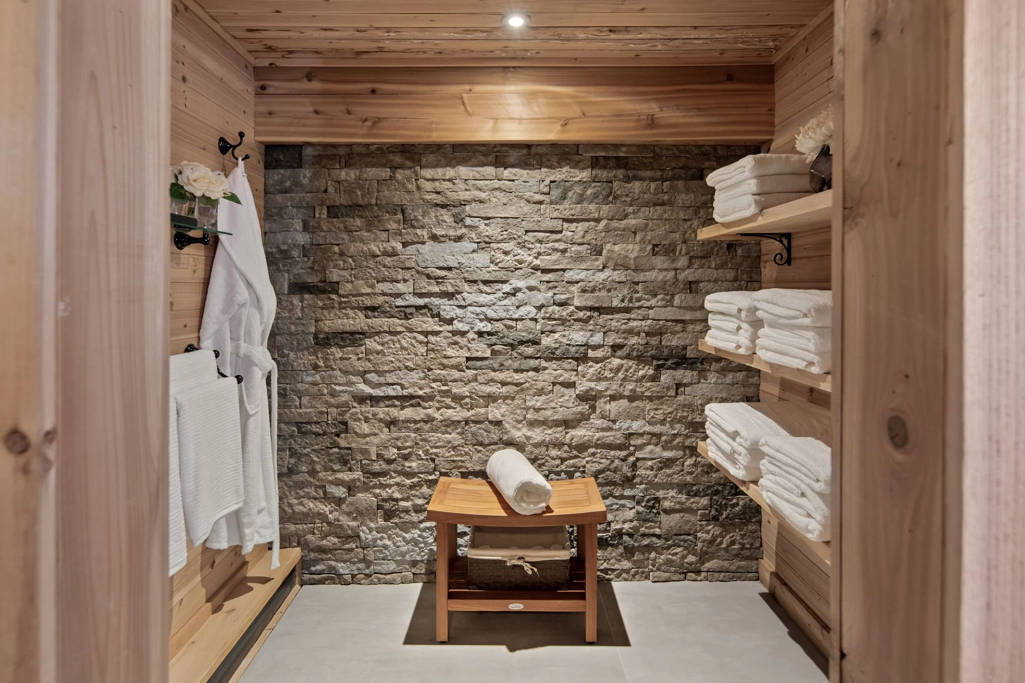 A cozy spa or wellness room with wooden walls and shelves holding neatly folded white towels. The back wall has a textured stone finish, and a small wooden table with a rolled towel on top is centered in the room. There are flowers and hooks on the l