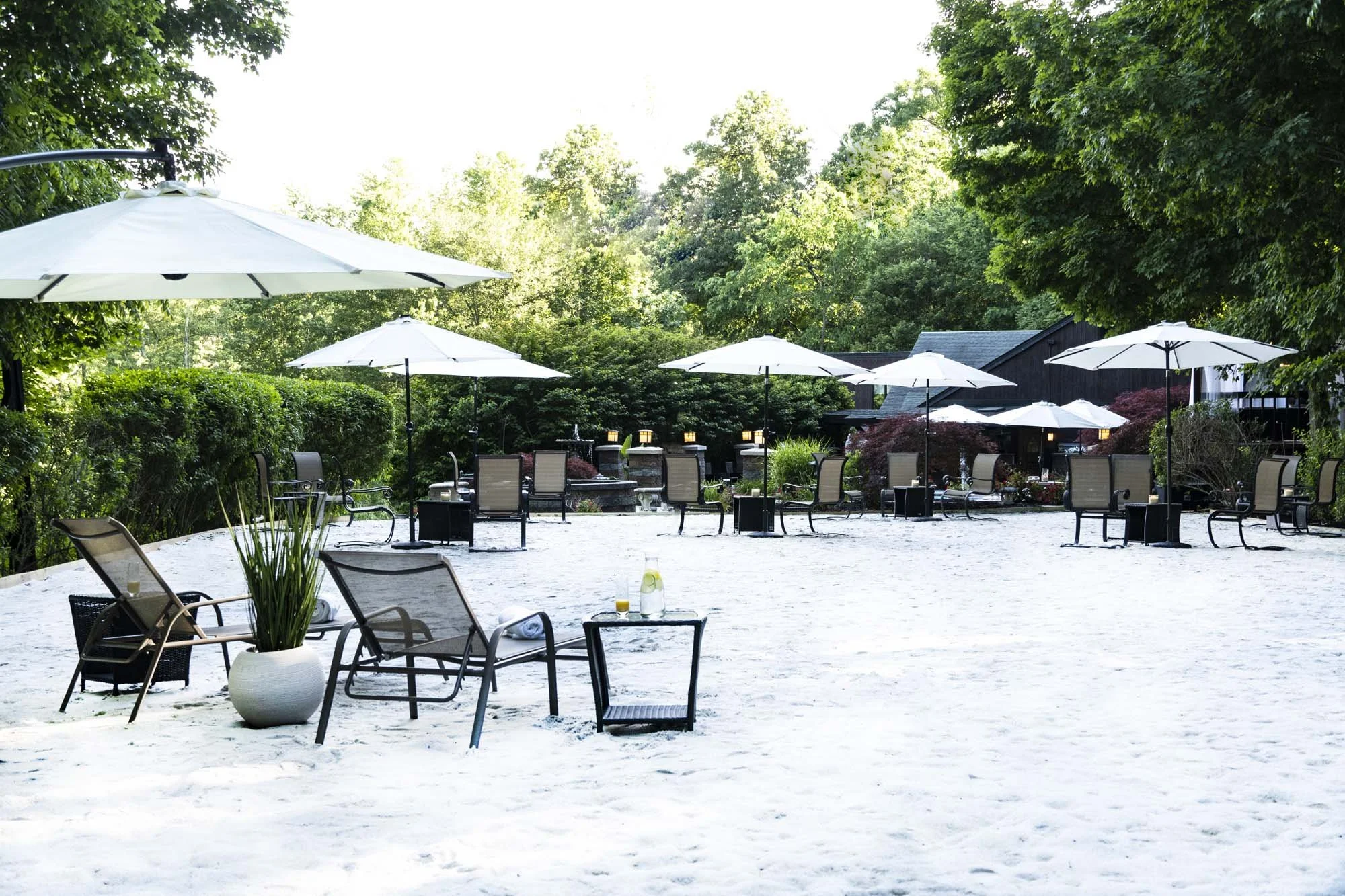 An outdoor patio area with white sand ground, several white umbrellas, black mesh chairs, small tables, and some set-up for drinks, surrounded by lush green trees and a dark building in the background.