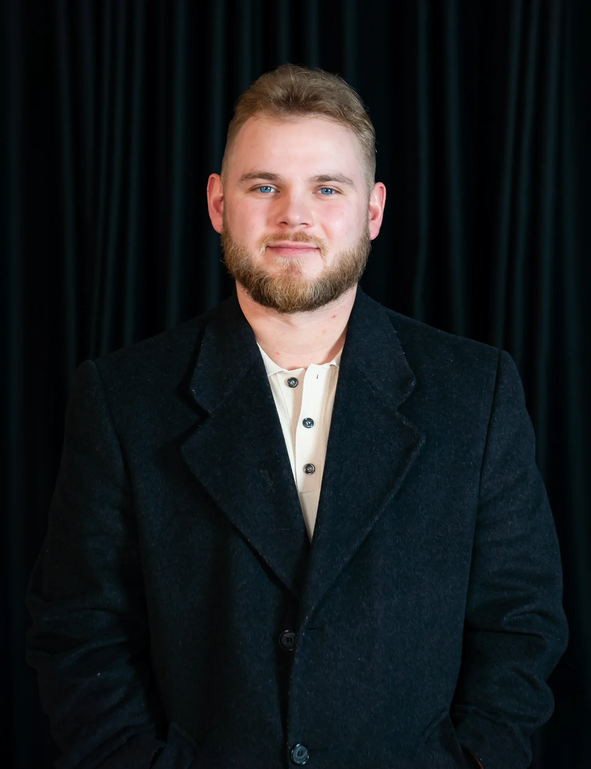 A young man with light brown hair, a beard, and blue eyes, wearing a black coat over a cream-colored button-up shirt, standing in front of a dark curtain background.