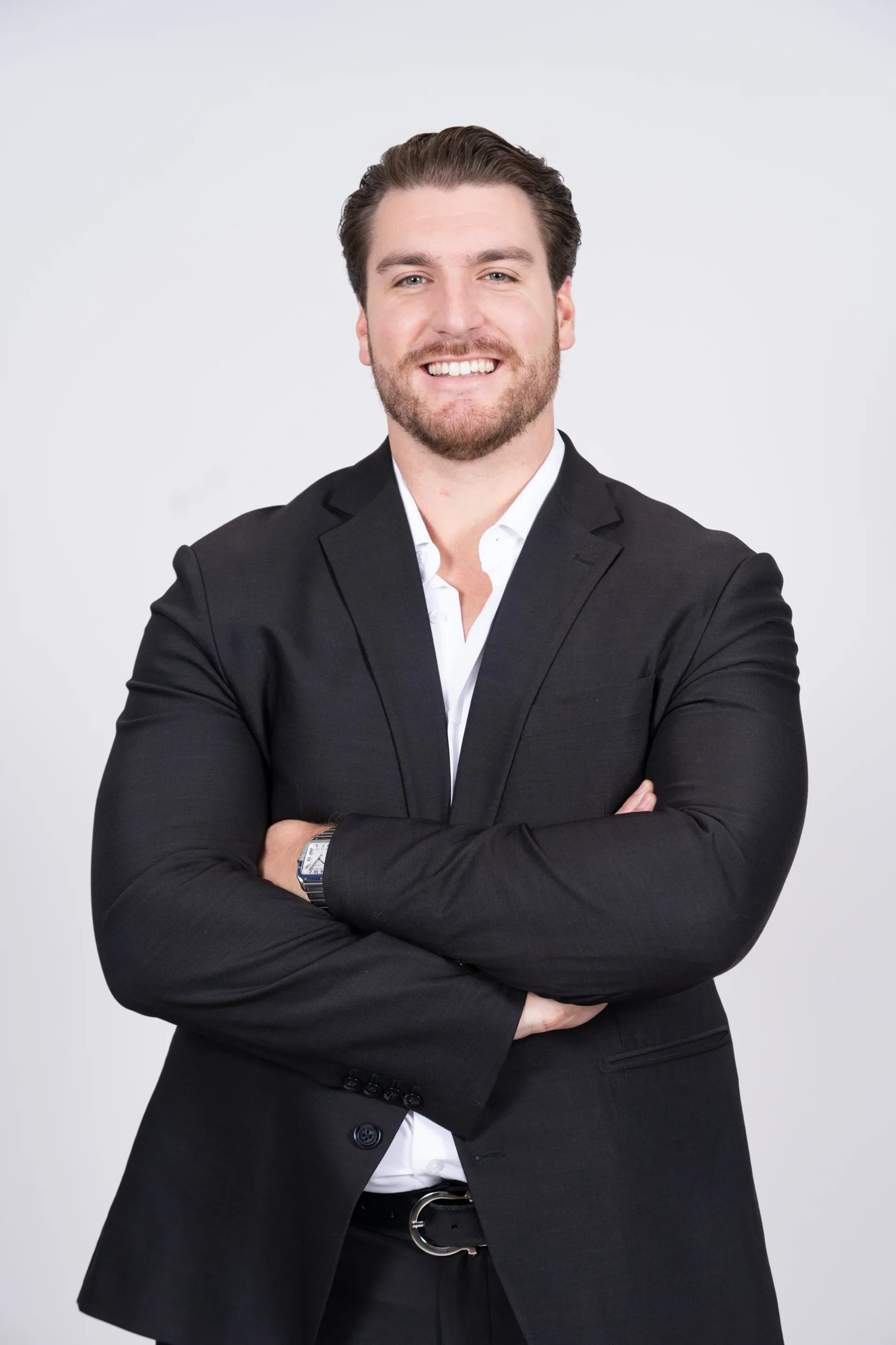 A man with brown hair and a beard, wearing a black suit with a white dress shirt, smiling with crossed arms against a plain white background.
