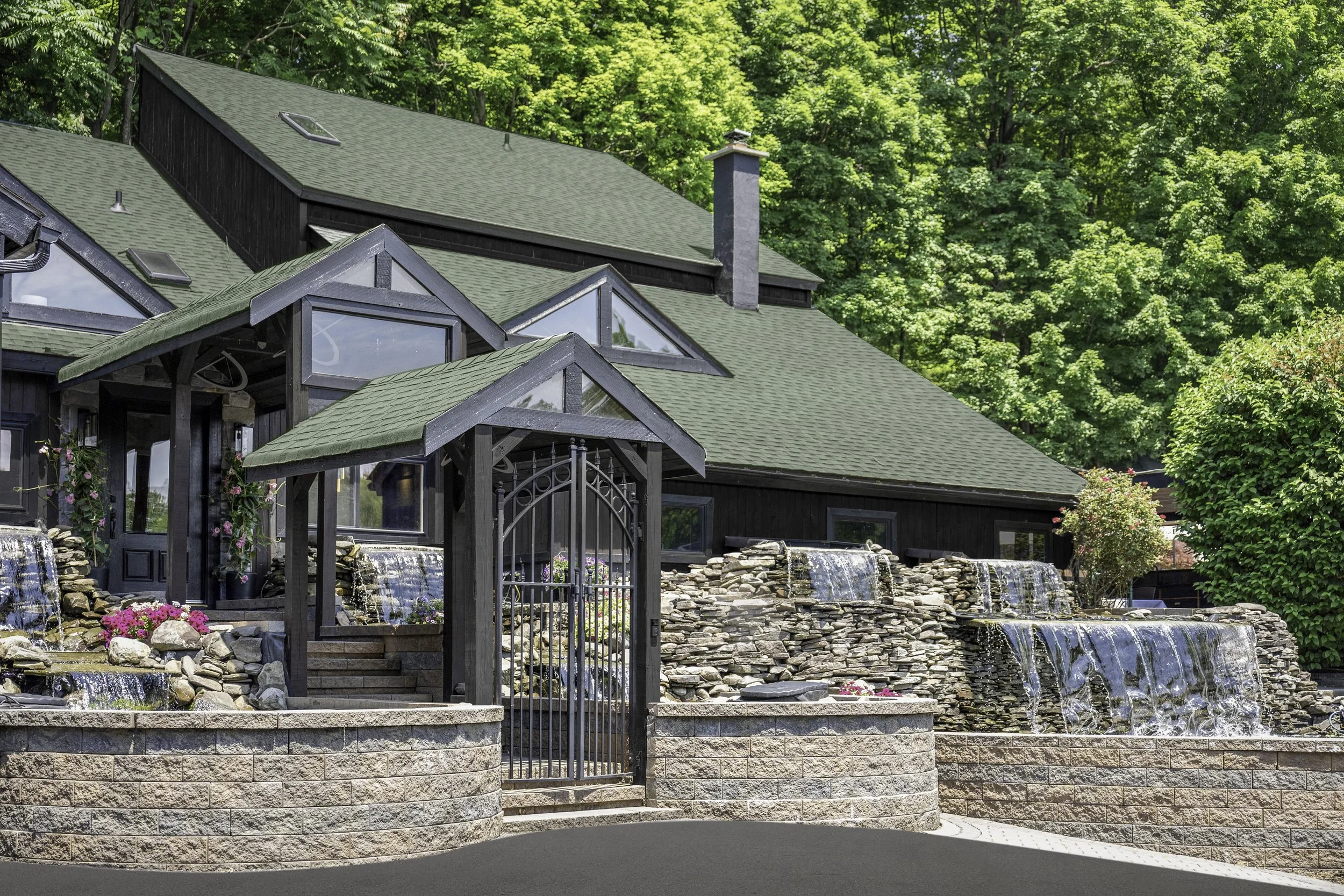 A house with a green roof, multiple chimneys, and large windows is surrounded by lush green trees. In front, there is a decorative stone wall, a fountain with water cascading over rocks, and a black metal gate.