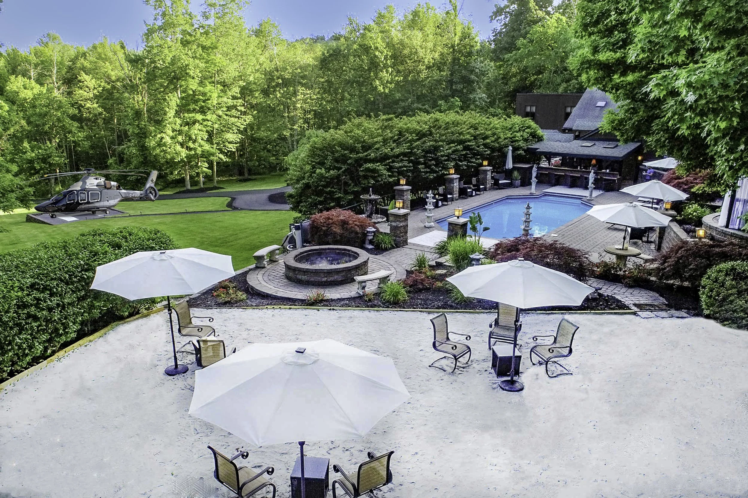 Outdoor backyard area with a swimming pool, patio seating with umbrellas, garden sculptures, and a helicopter displayed on a lawn surrounded by lush green trees.