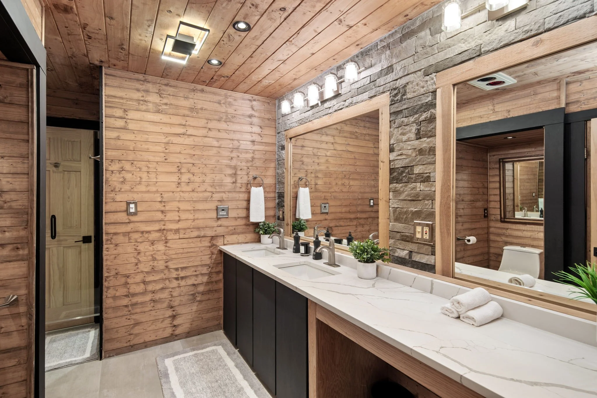 A modern bathroom with wood-paneled walls and ceiling, stone accents, a double sink vanity with a marble countertop, and a large mirror. There are two white towels on the counter and a small potted plant. The lighting fixtures include a row of six gl