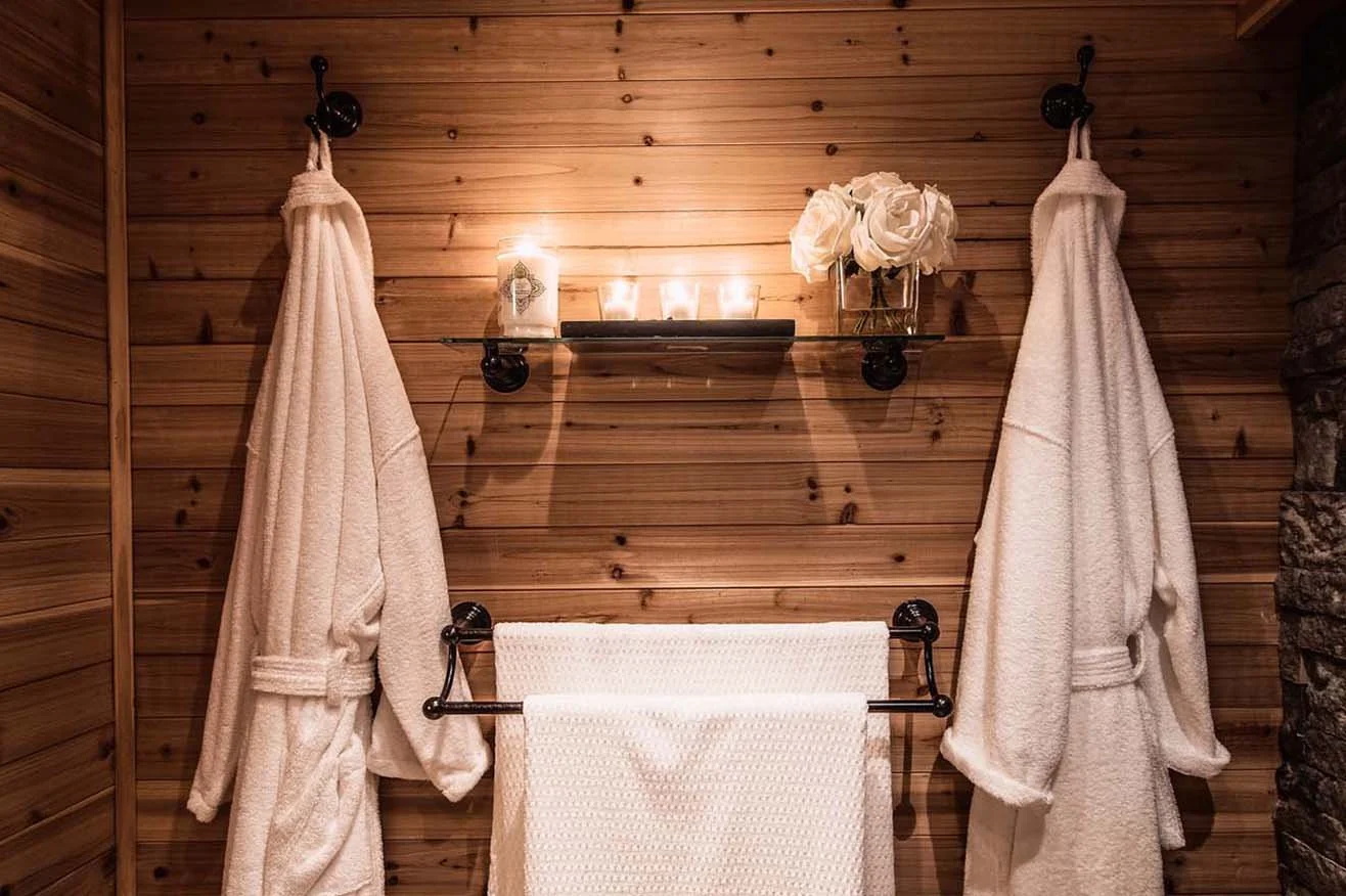 A rustic bathroom with wooden walls, two white bathrobes hanging on hooks, a glass shelf with candles and a vase of white roses, and a towel rack with a white towel.
