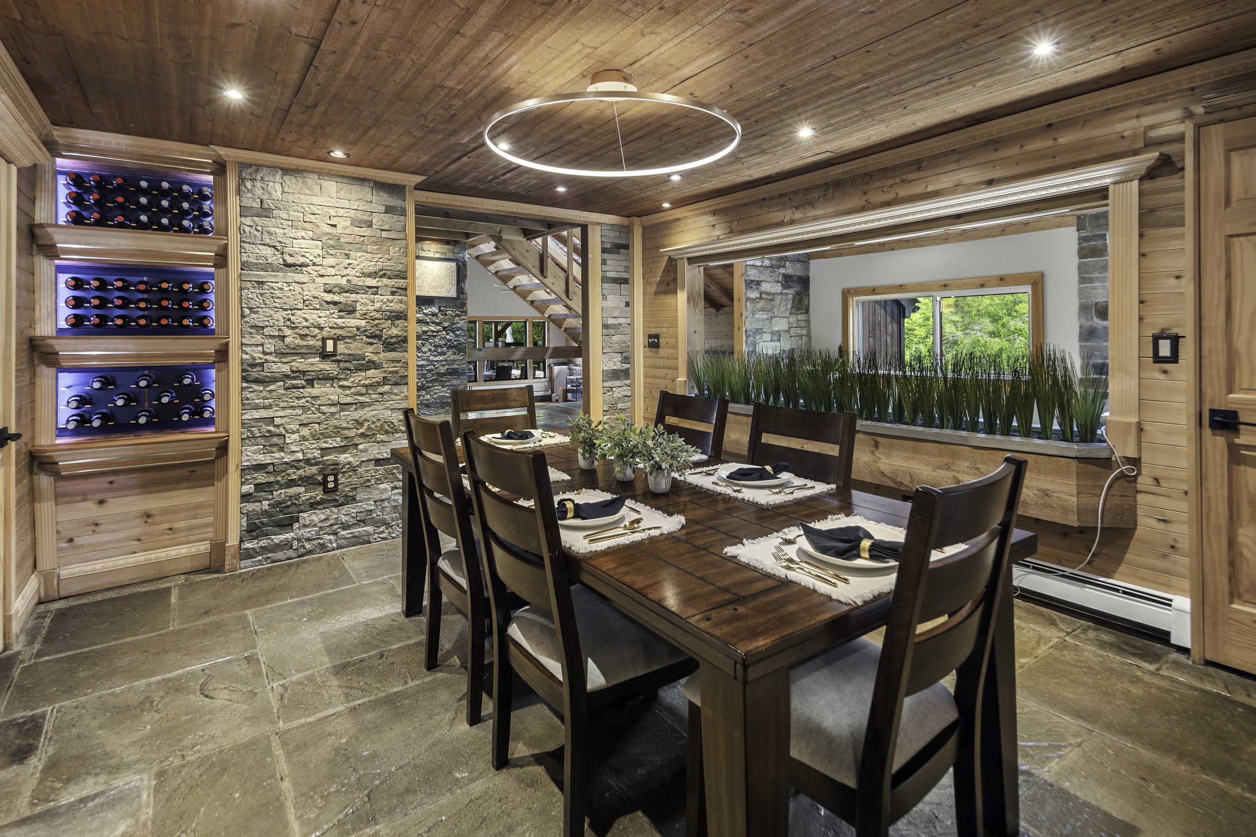 A cozy dining room with a wooden table set for dinner with plates, napkins, and utensils. The space features wood-paneled ceiling, stone and wood walls, and a large window with green foliage outside. There is a modern circular ceiling light and built