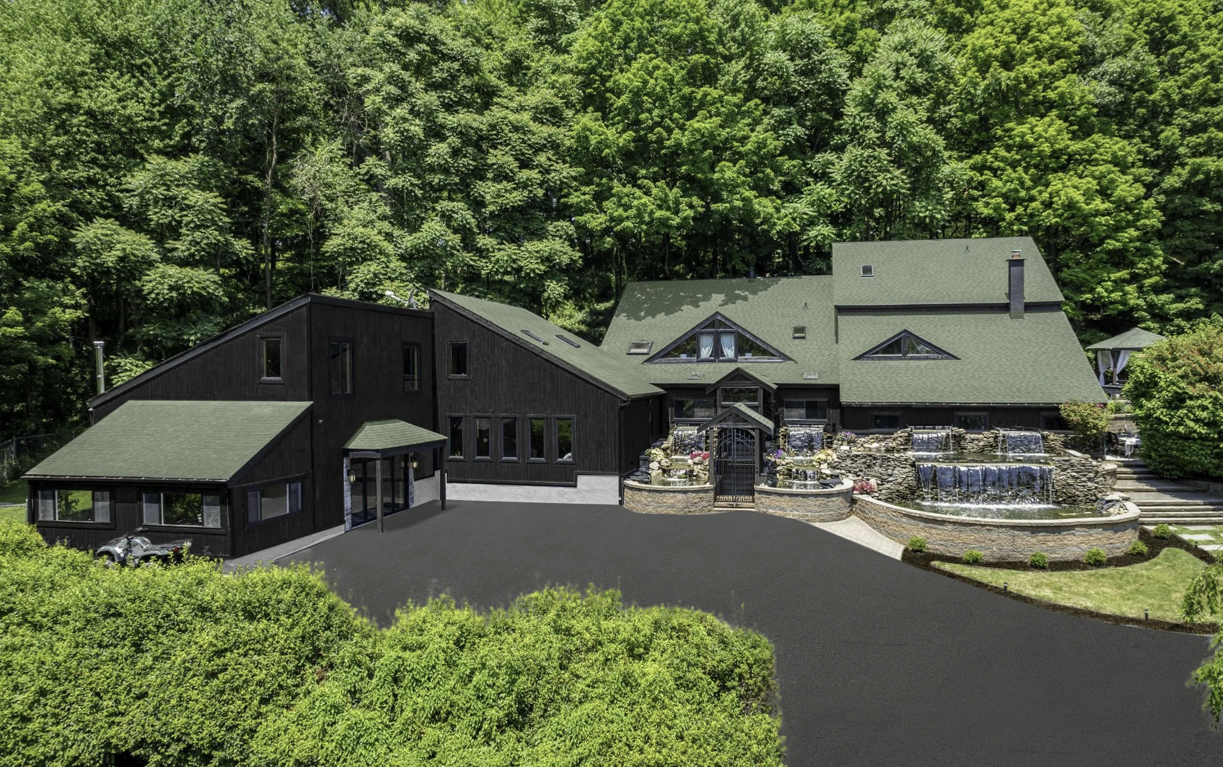 A modern house with a green roof, black exterior walls, and large windows is surrounded by lush green trees and a landscaped yard with a waterfall feature.