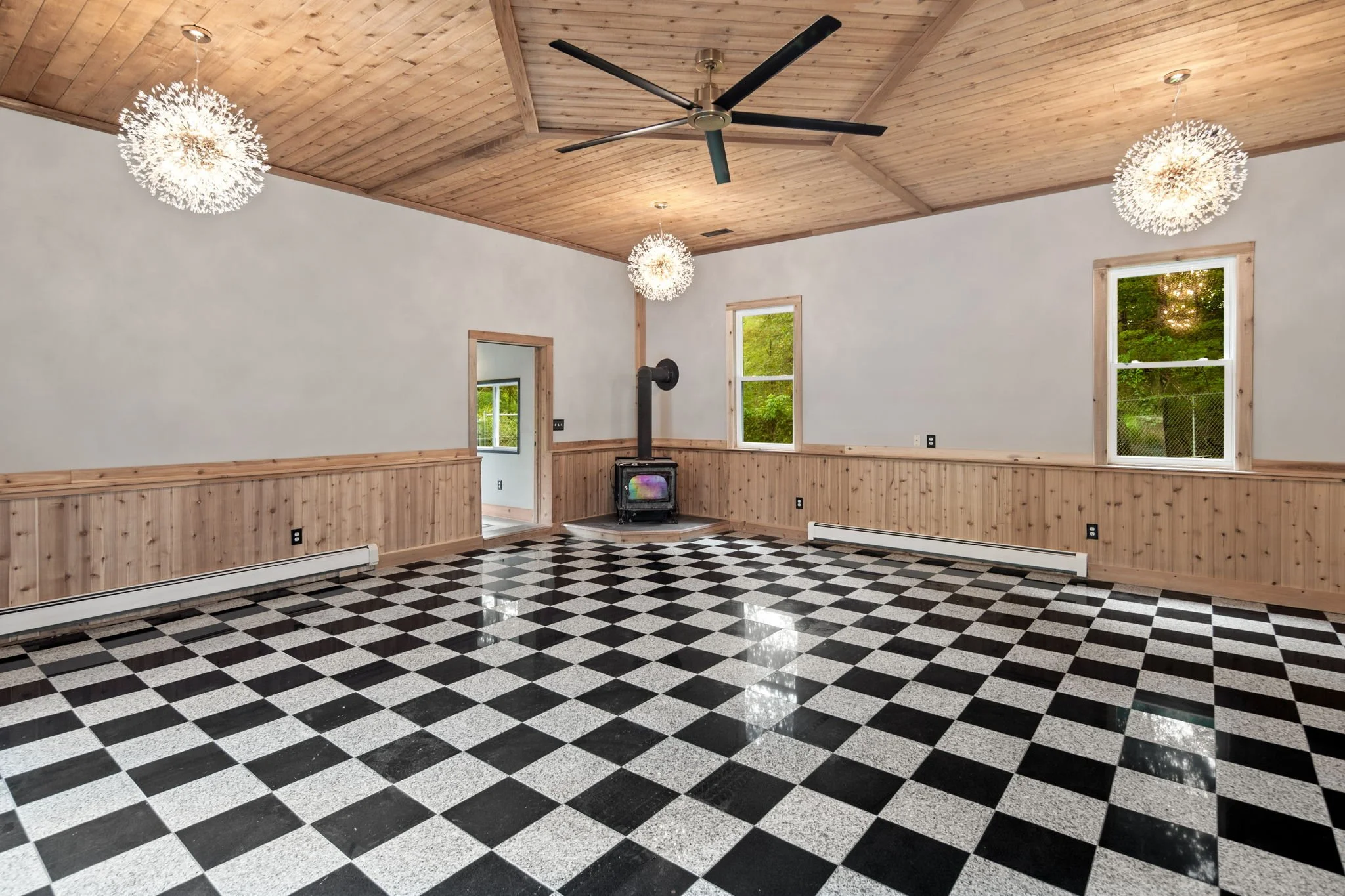 Interior of a room with a checkered black and white floor, wooden wainscoting, and a wooden ceiling with three hanging light fixtures, two windows with views of trees, a black ceiling fan, and a wood stove.