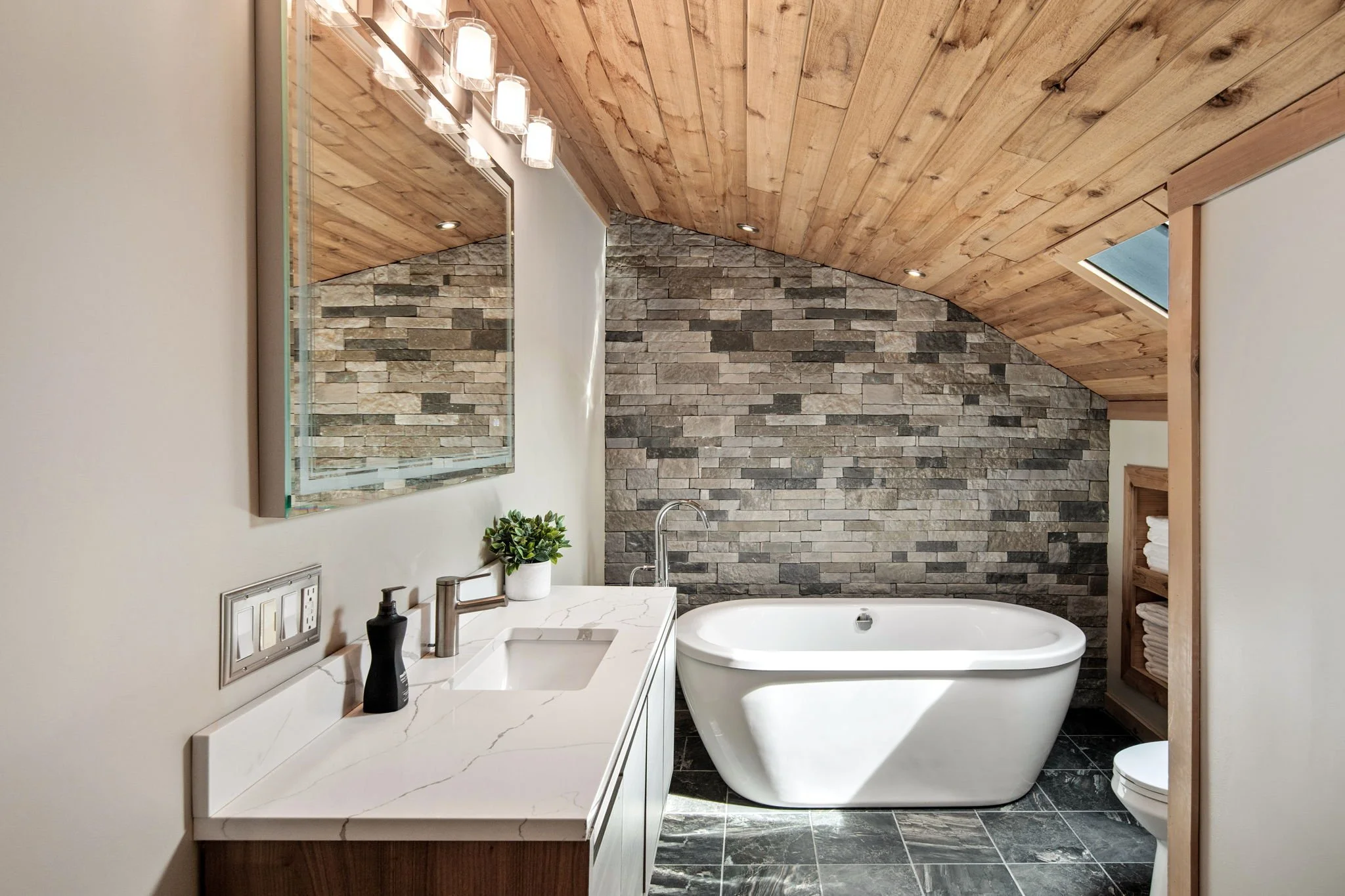 Modern bathroom with a white vanity, a rectangular mirror, black soap dispenser, small potted plant, freestanding bathtub, stone accent wall, wood-paneled ceiling, and skylight.