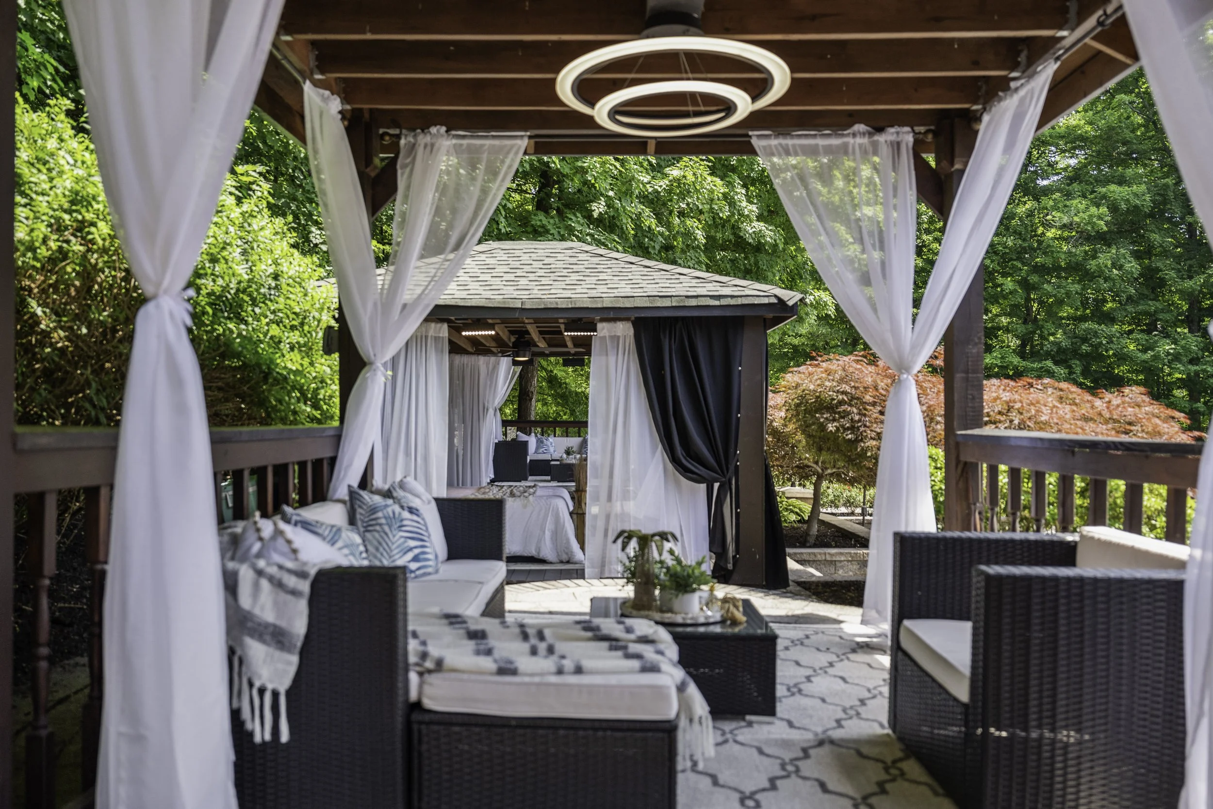 A covered outdoor patio with white curtains tied back, black wicker furniture with patterned cushions, and a modern circular chandelier, set among lush green trees and foliage.