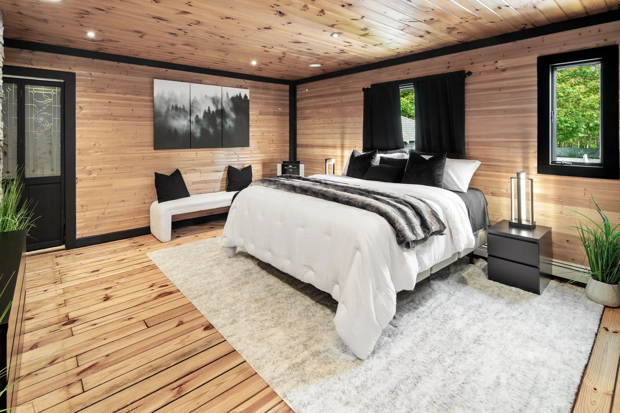 A cozy bedroom with wooden paneled walls and ceiling, a large bed with white bedding and black and gray pillows, a striped throw blanket, a beige bench with black pillows at the foot of the bed, two black nightstands with modern lamps, black-framed w