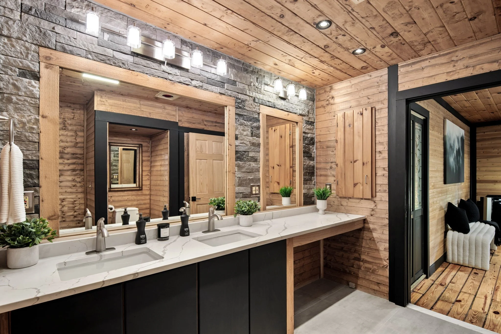 A rustic bathroom with wood-paneled walls and ceiling, a large mirror above a white marble countertop with two sinks, and various plants and toiletries on the counter.