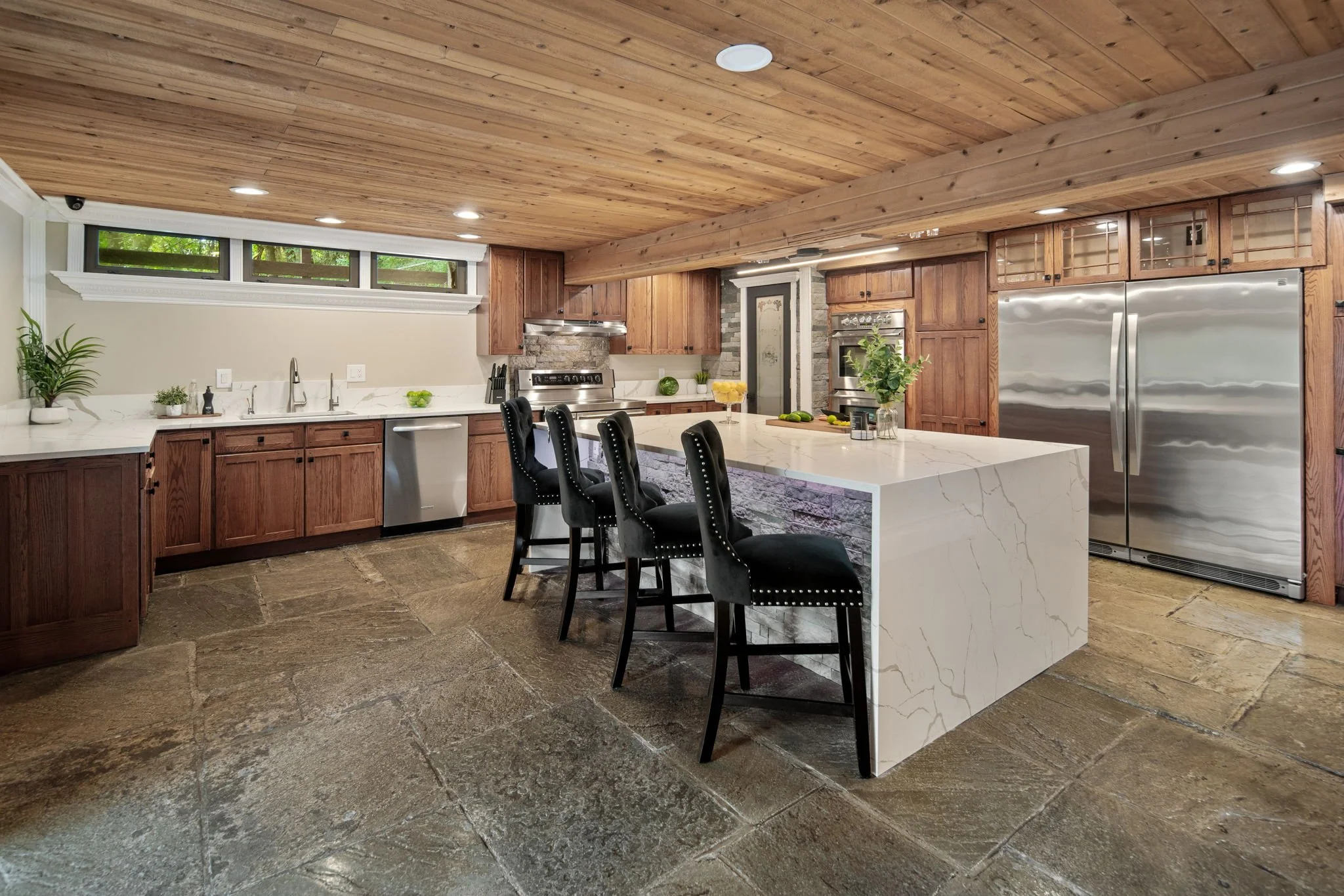 Modern kitchen with wooden cabinets, marble island, and stainless steel appliances, illuminated by recessed ceiling lights, with a wooden plank ceiling and stone tile flooring.