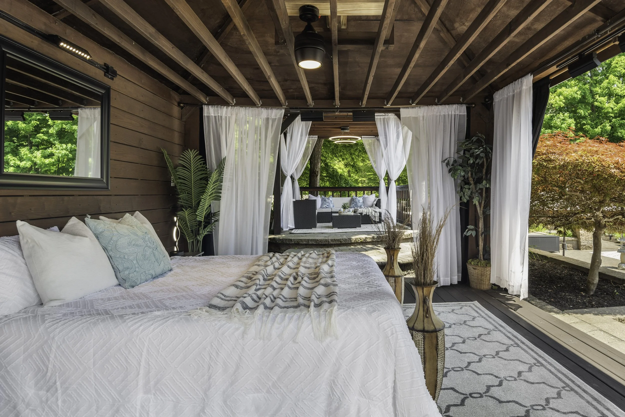 A cozy outdoor patio area with white curtains, green plants, and wicker furniture, surrounded by lush green trees.