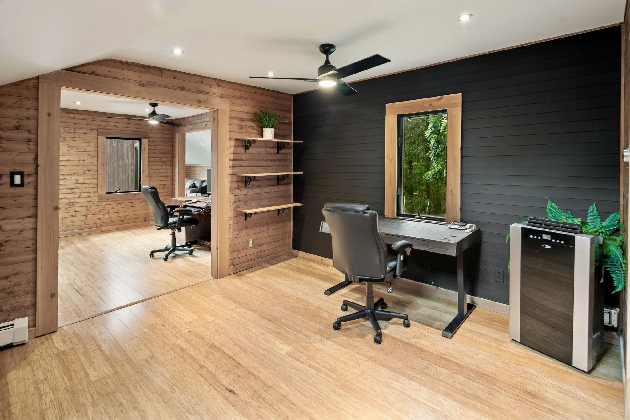 Home office with wooden walls and flooring, black and gray furniture, large windows with green view, ceiling fans, and potted plants.