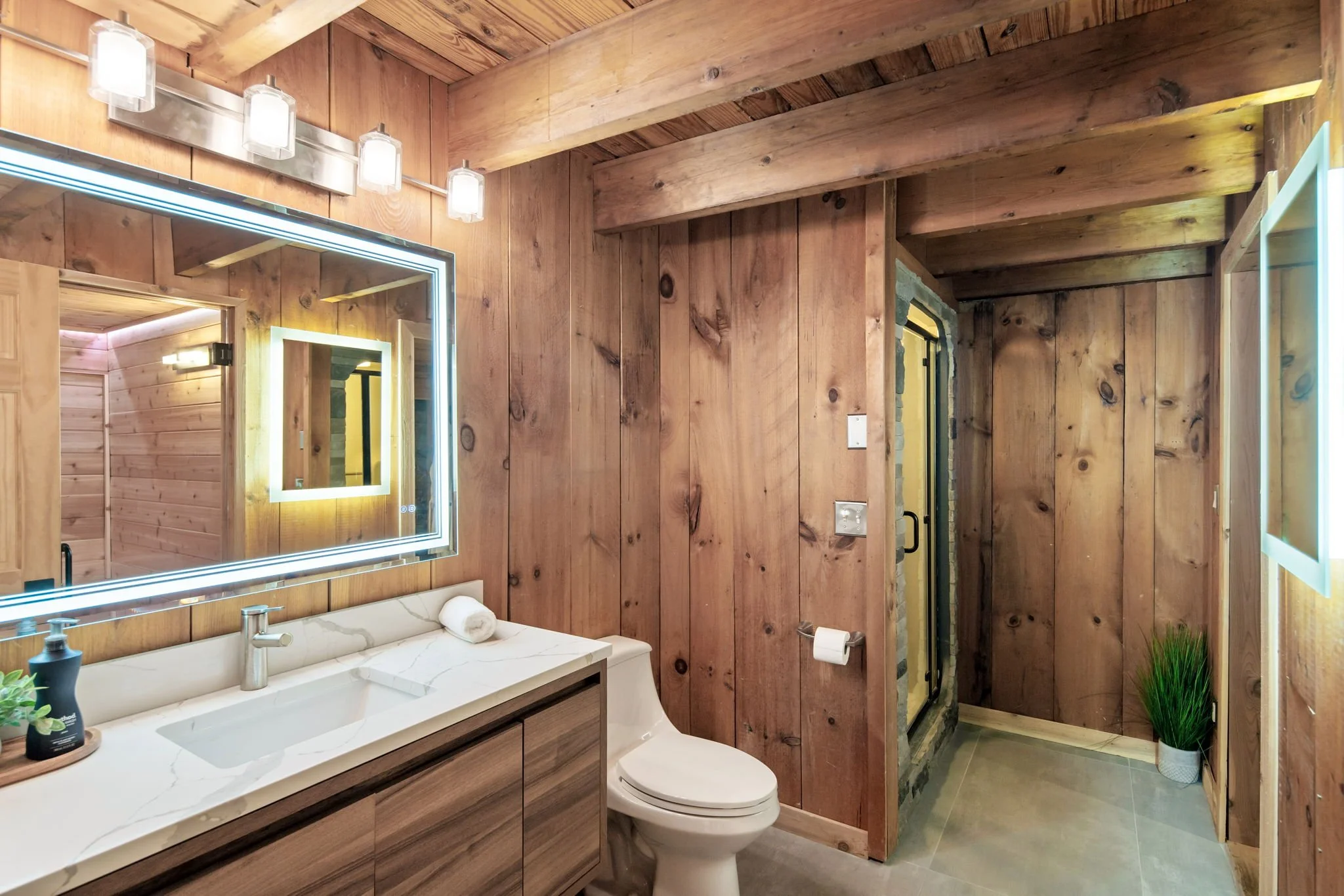 A rustic wooden bathroom featuring a marble countertop with a built-in sink, illuminated rectangular mirror with LED lights, modern faucet, a toilet, and a shower area enclosed with a glass door. There are hanging lights above the mirror, a small pla