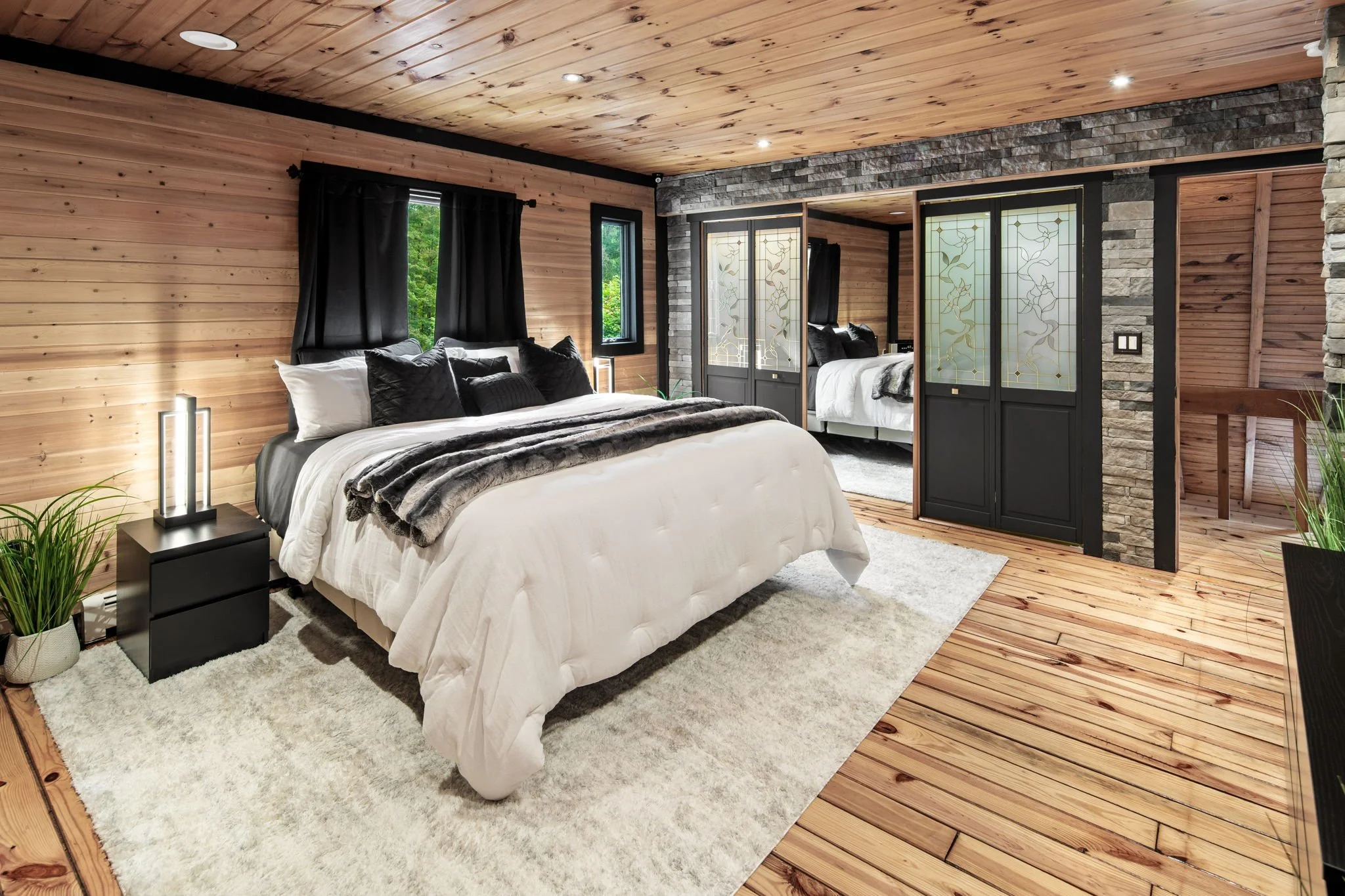 A bedroom with wood-paneled walls and ceiling, a large bed with white and black bedding, black curtains, a small bedside table with a lamp, and a sliding door with decorative frosted glass panels. There are two windows showing green trees outside.