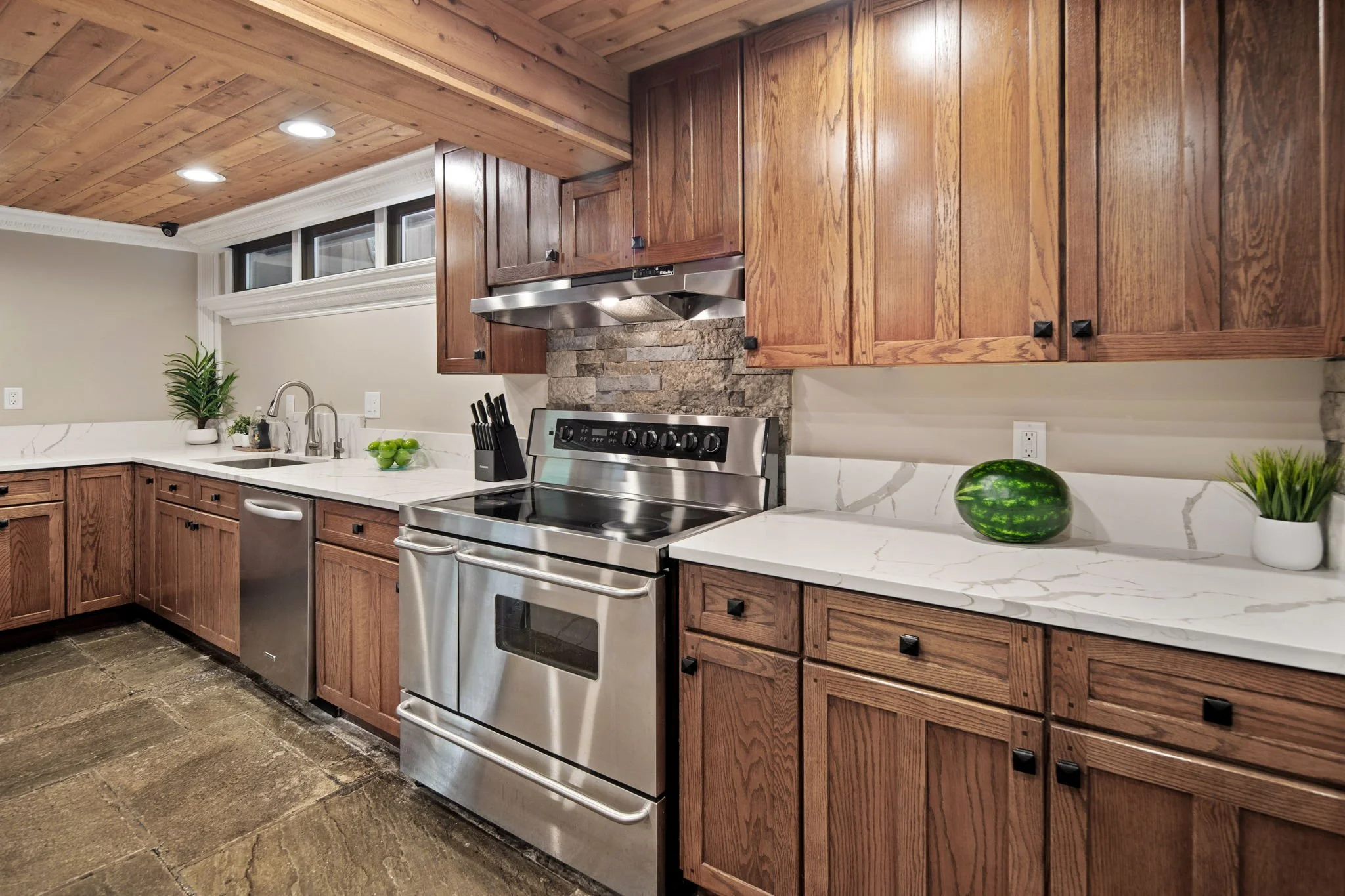 A modern kitchen with wooden cabinets, a marble countertop, a stainless steel stove, a water watermelon on the counter, and a potted plant near the sink. Recessed lighting is visible in the wooden ceiling.
