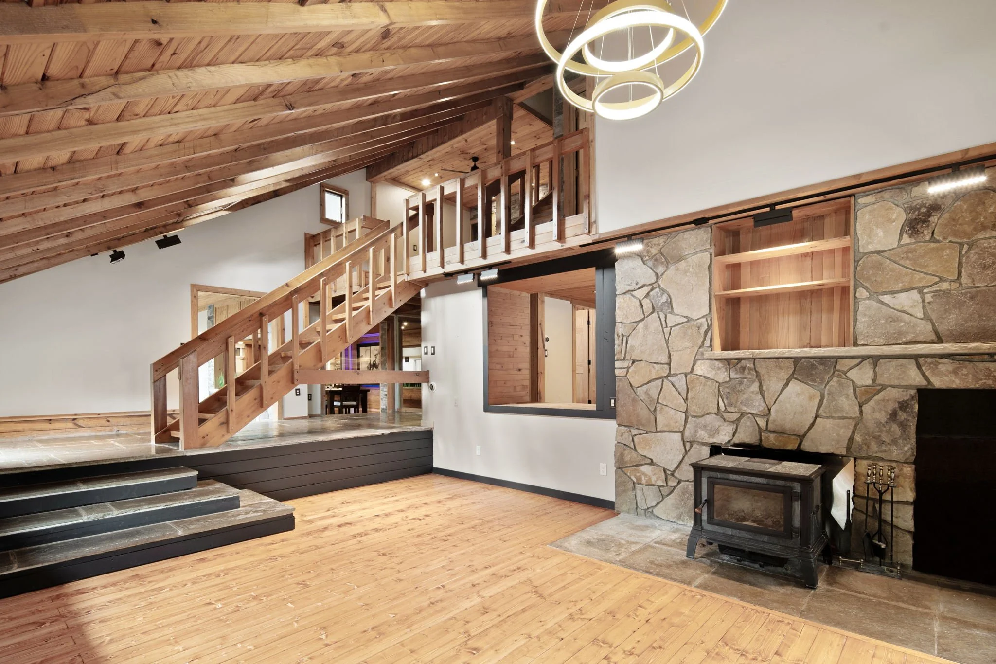 Interior of a modern home with wooden ceiling, stairs, and a stone fireplace with a wood stove. There is a circular chandelier hanging from the ceiling.