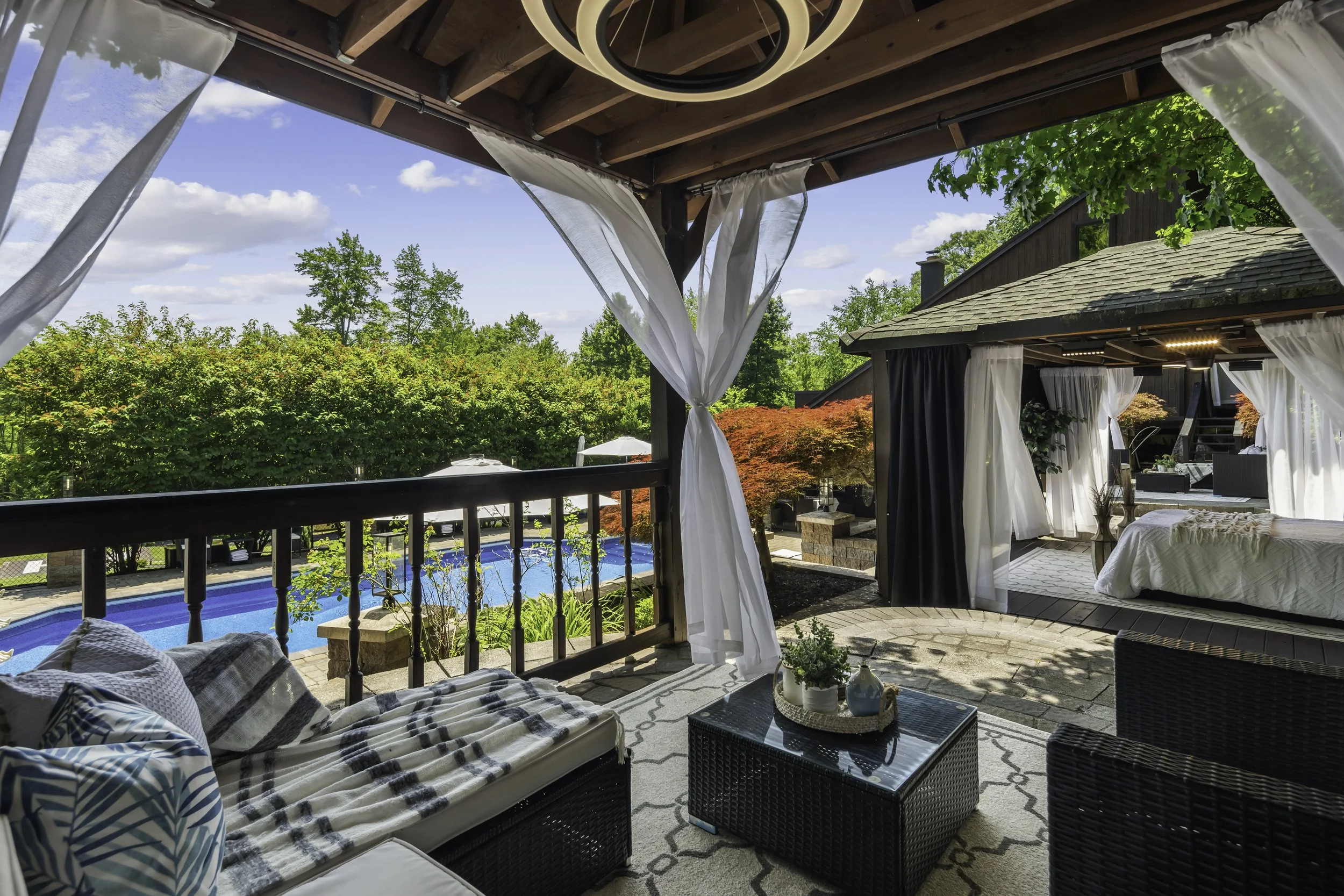 A covered outdoor patio with seating furniture, including a sofa and wicker chairs, overlooking a backyard with a swimming pool, lush greenery, and trees under a blue sky with scattered clouds.