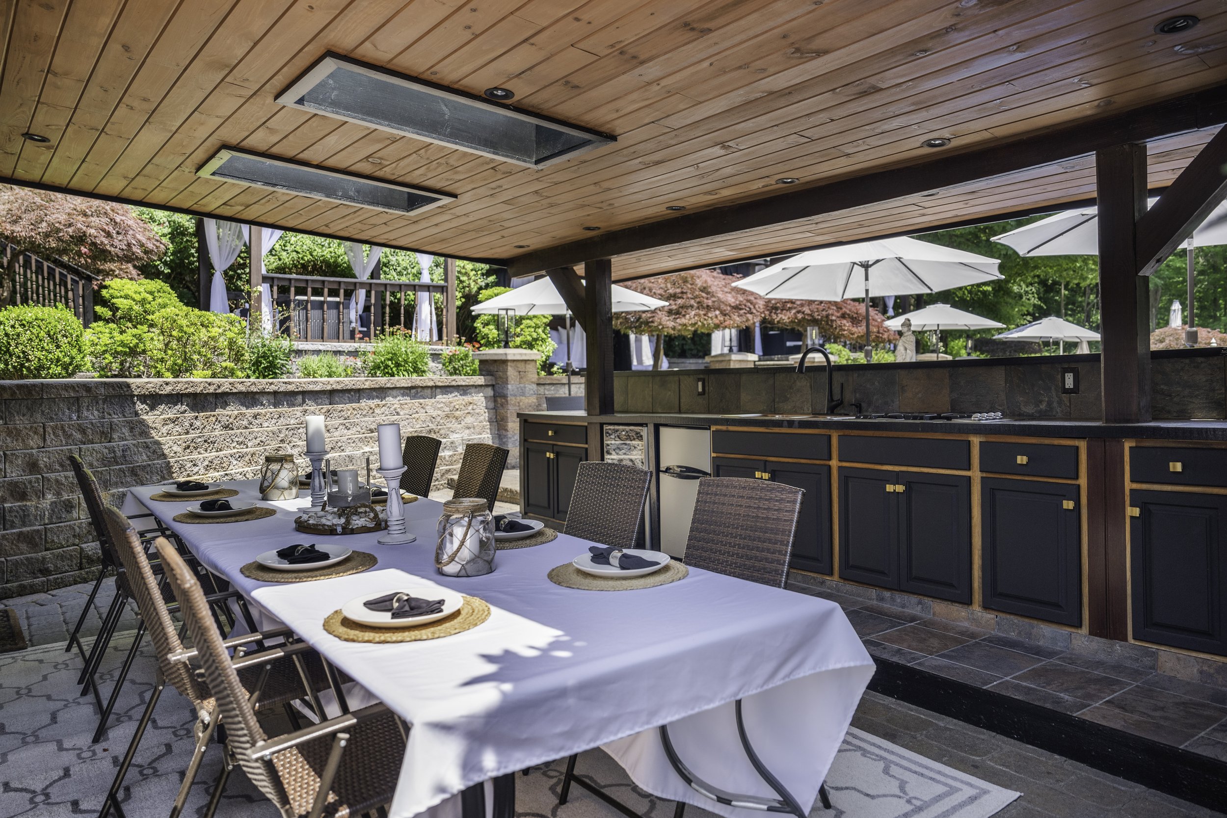 Outdoor dining area with a long table set for a meal, surrounded by chairs, under a wooden ceiling with recessed lighting, overlooking a lush garden with trees, umbrellas, and a deck.