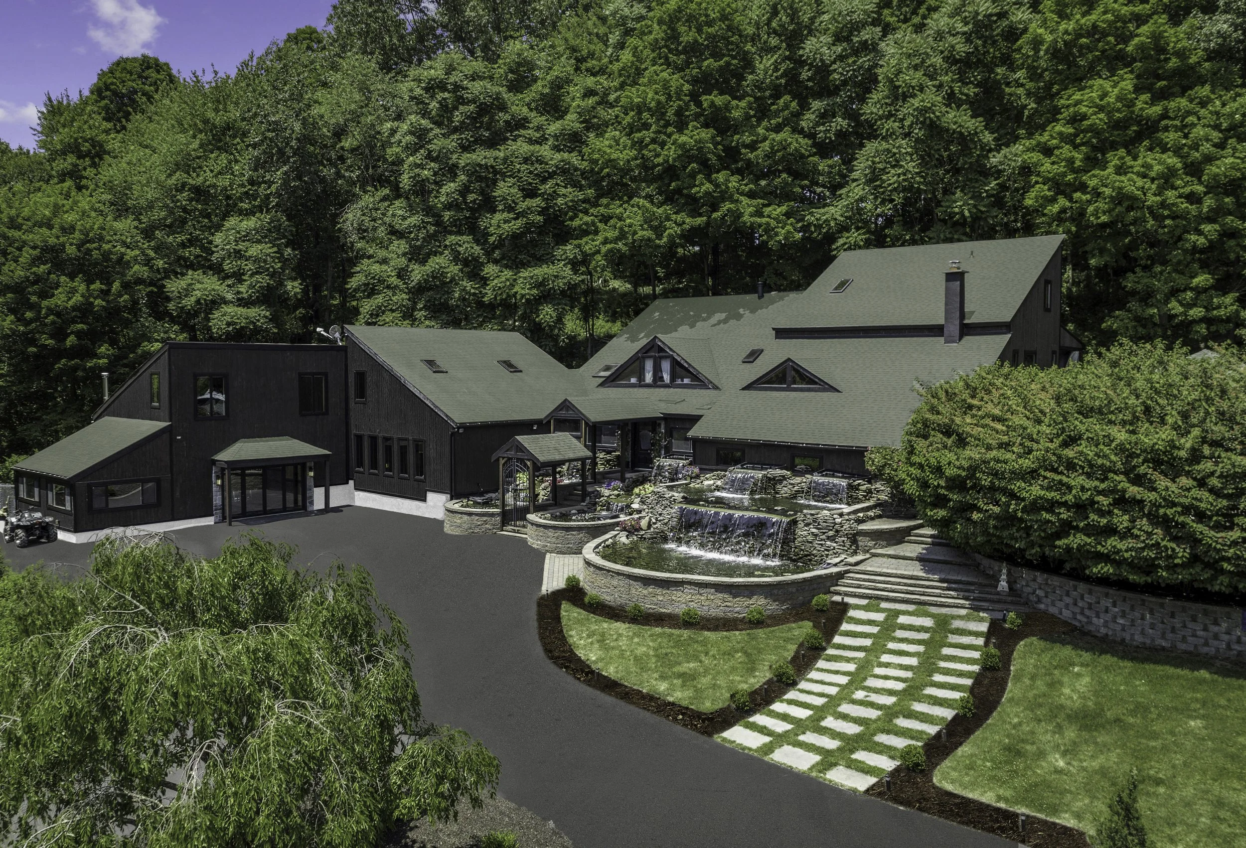 A modern house with a green roof surrounded by lush green trees and well-maintained landscaping, including a small pond with cascading waterfalls, a stone pathway, and a paved driveway.