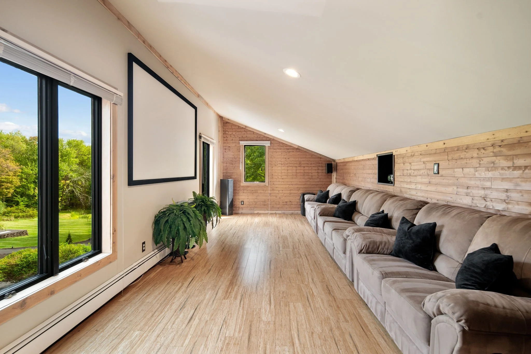 A spacious living room with large windows showing a lush green landscape outside. The room has a high, sloped ceiling with recessed lighting, wooden paneling on one wall, and light hardwood flooring. A beige sectional sofa with black throw pillows is positioned along the wooden wall, and there are potted plants near the windows.