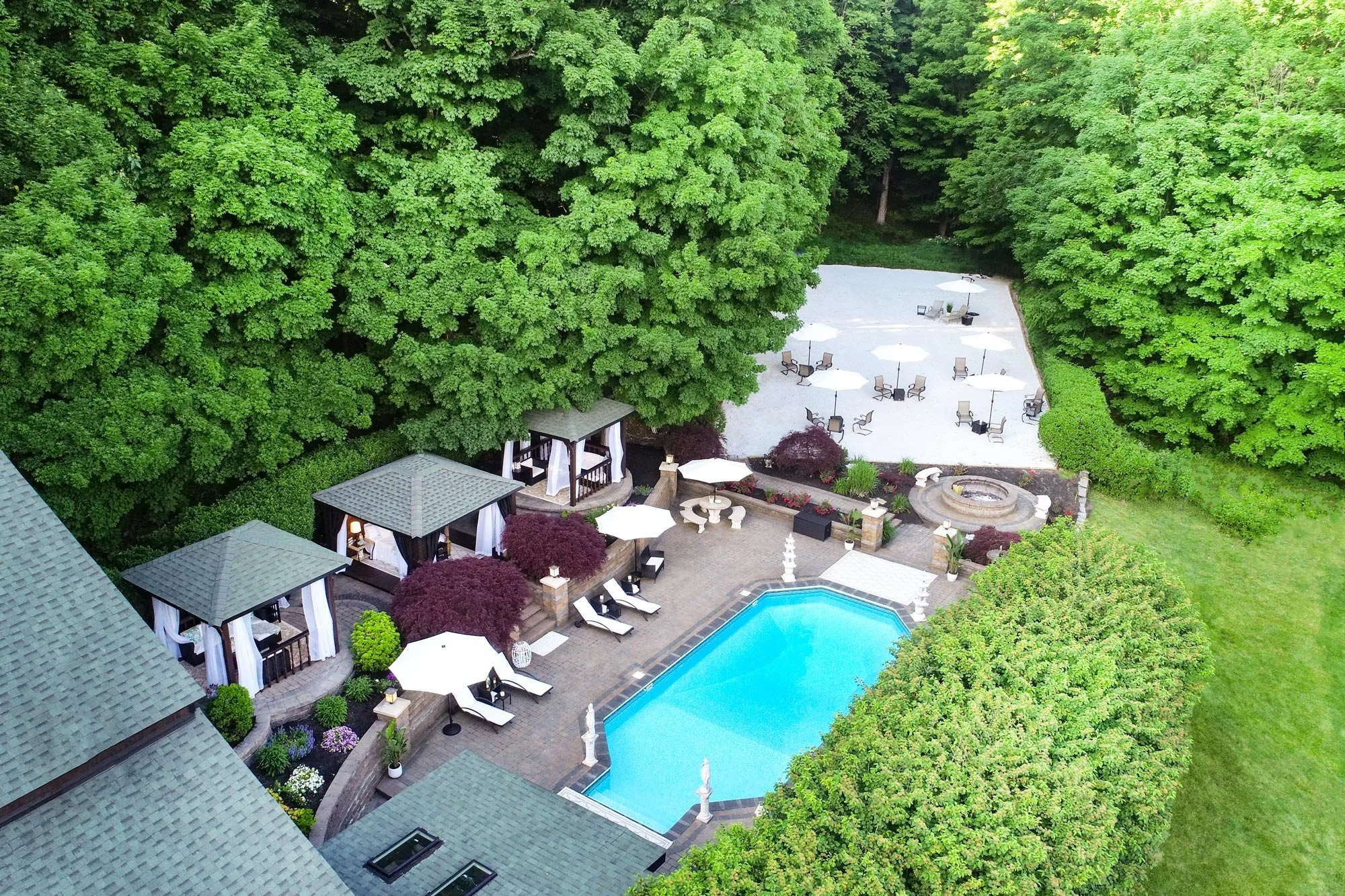 An aerial view of a backyard with a swimming pool, surrounded by lush green trees, outdoor patio furniture, umbrellas, and a small patio with a fountain.