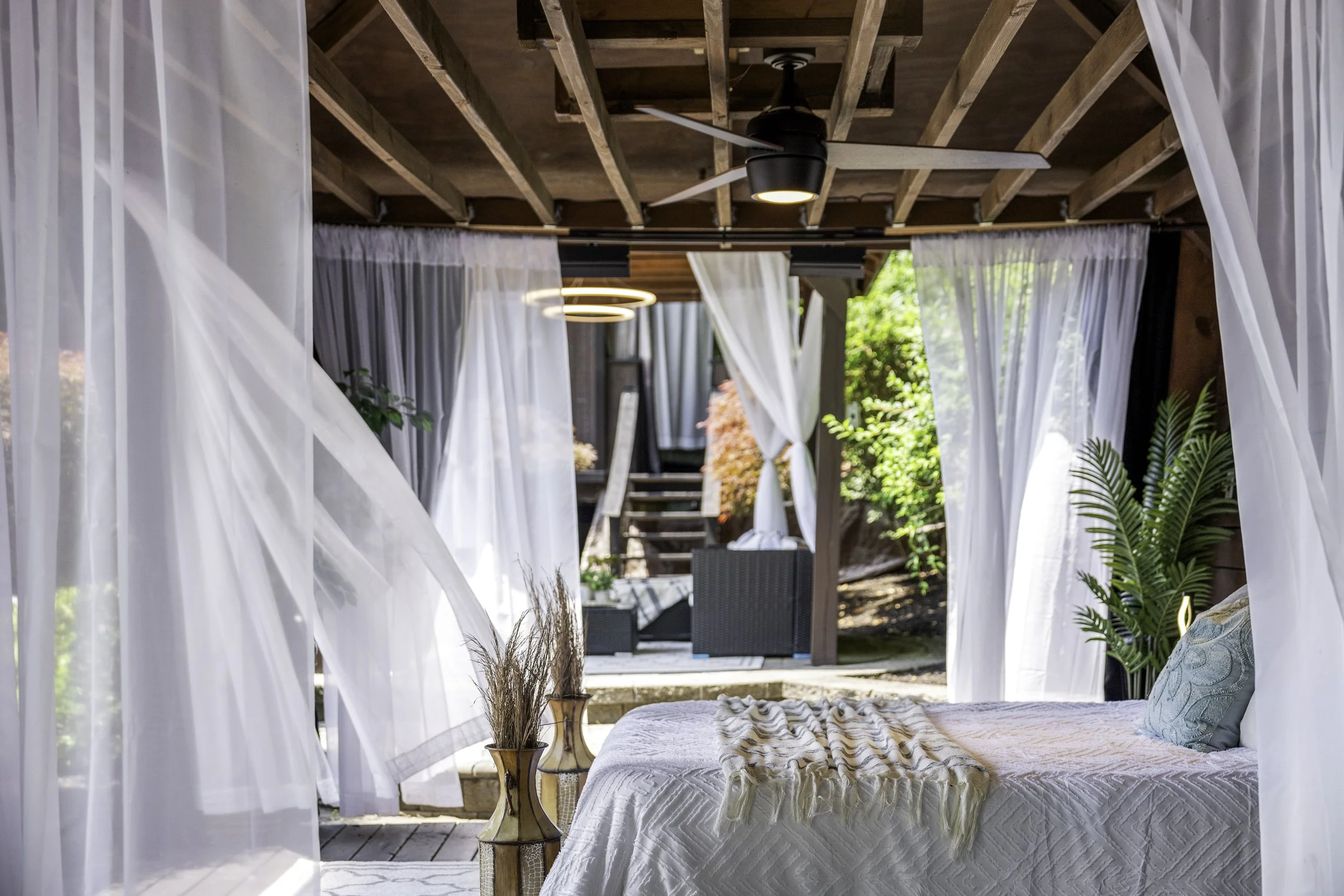 A bedroom with a canopy bed featuring white curtains and a textured bedspread. There's a small wooden side table with dried plants and a wooden chair. The room opens to an outdoor seating area with black wicker furniture, surrounded by lush greenery 