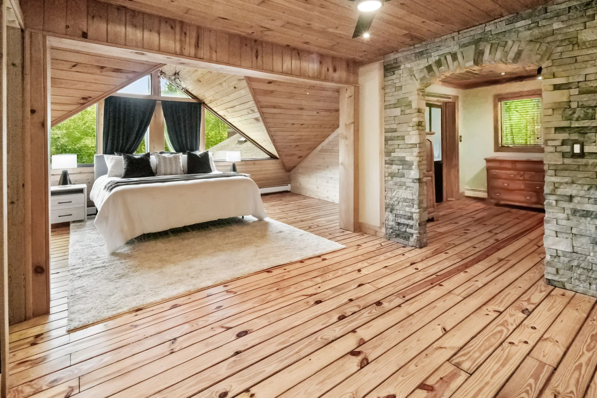 A cozy bedroom with wooden walls, ceiling, and floor. The bed has white bedding and black pillows, situated under an angled triangular window with dark curtains. A white nightstand with a lamp is beside the bed. The room features a partial stone wall