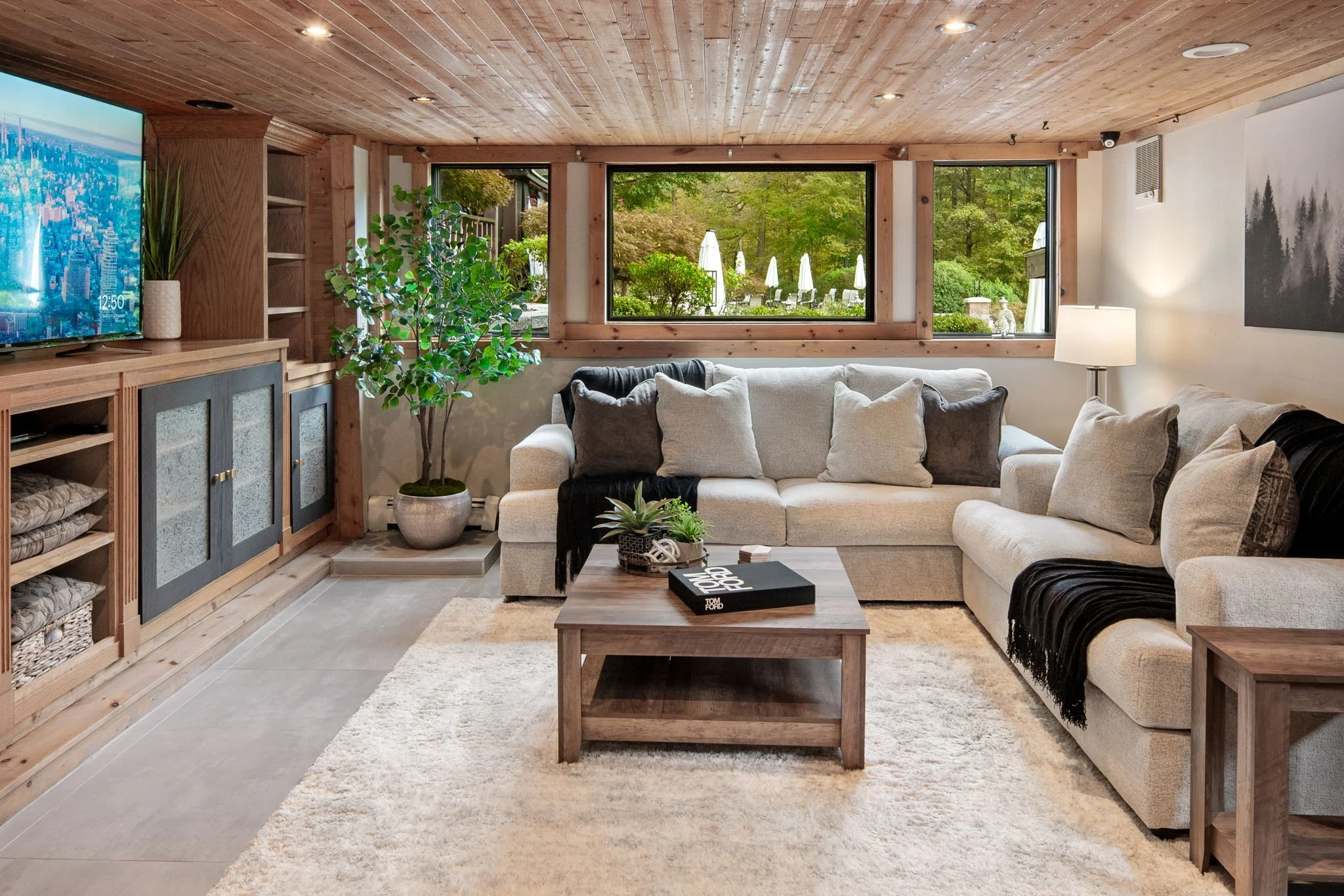 Living room with beige sofa, coffee table, wood-paneled ceiling, large window overlooking green outdoor space, TV on wooden cabinet, potted plant, floor lamp, art on wall, cozy and modern decor.