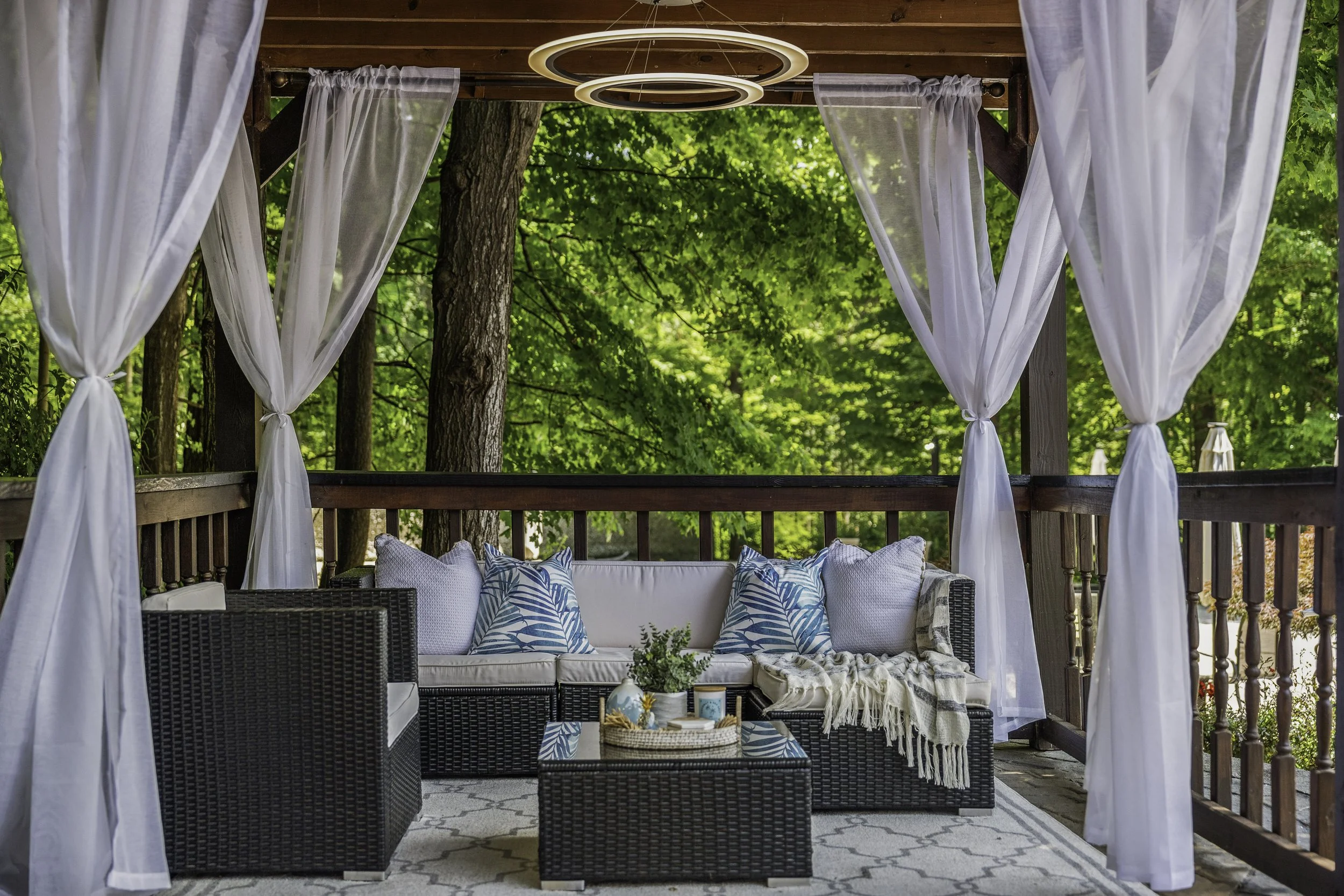 A screened porch with white curtains, a wicker couch with white cushions and blue leaf-patterned throw pillows, a black wicker coffee table with decor, and a lush green nature background.
