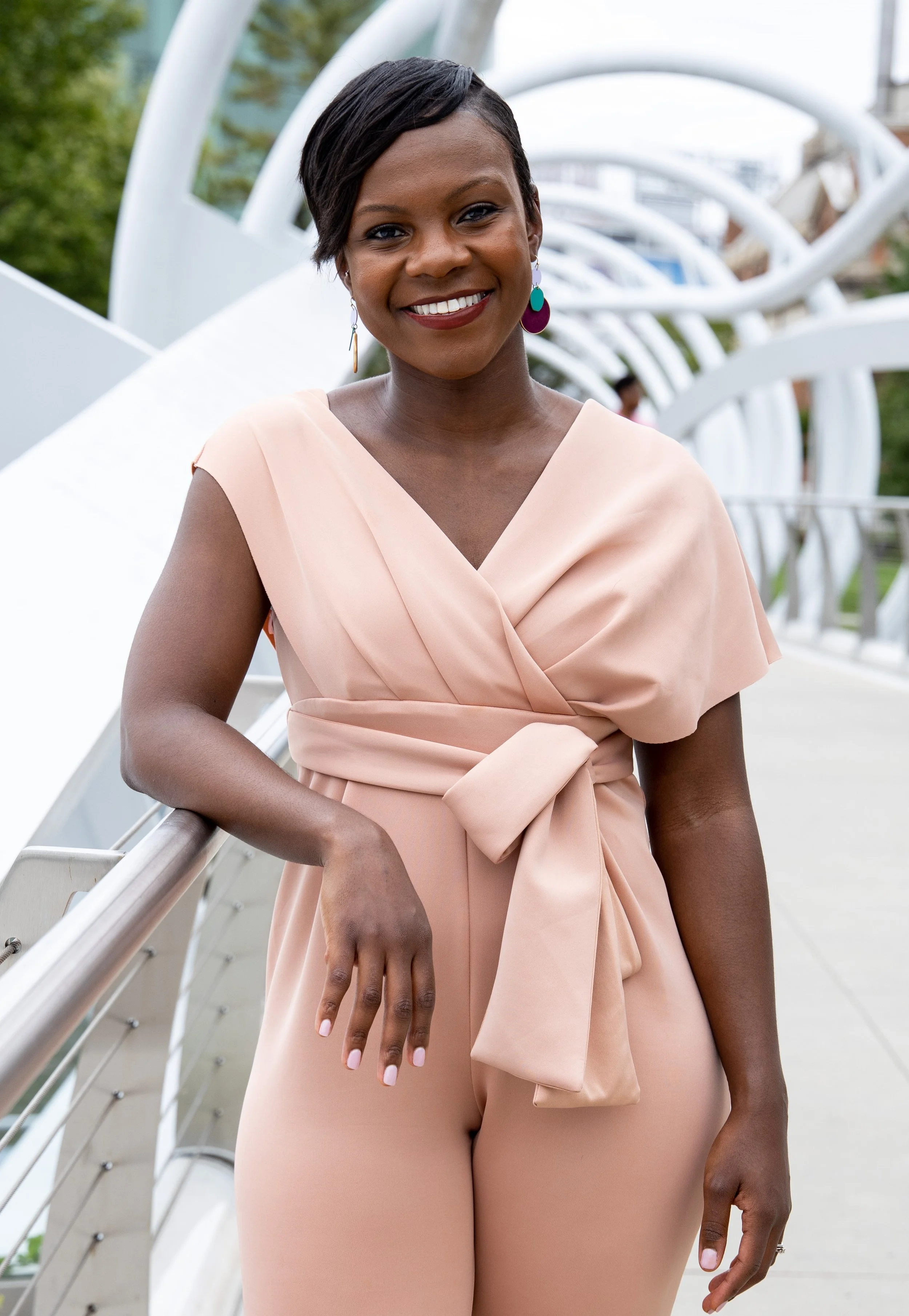 A smiling woman in a blush pink dress with short dark hair and colorful earrings, standing on a modern outdoor bridge or walkway with white curved structures.