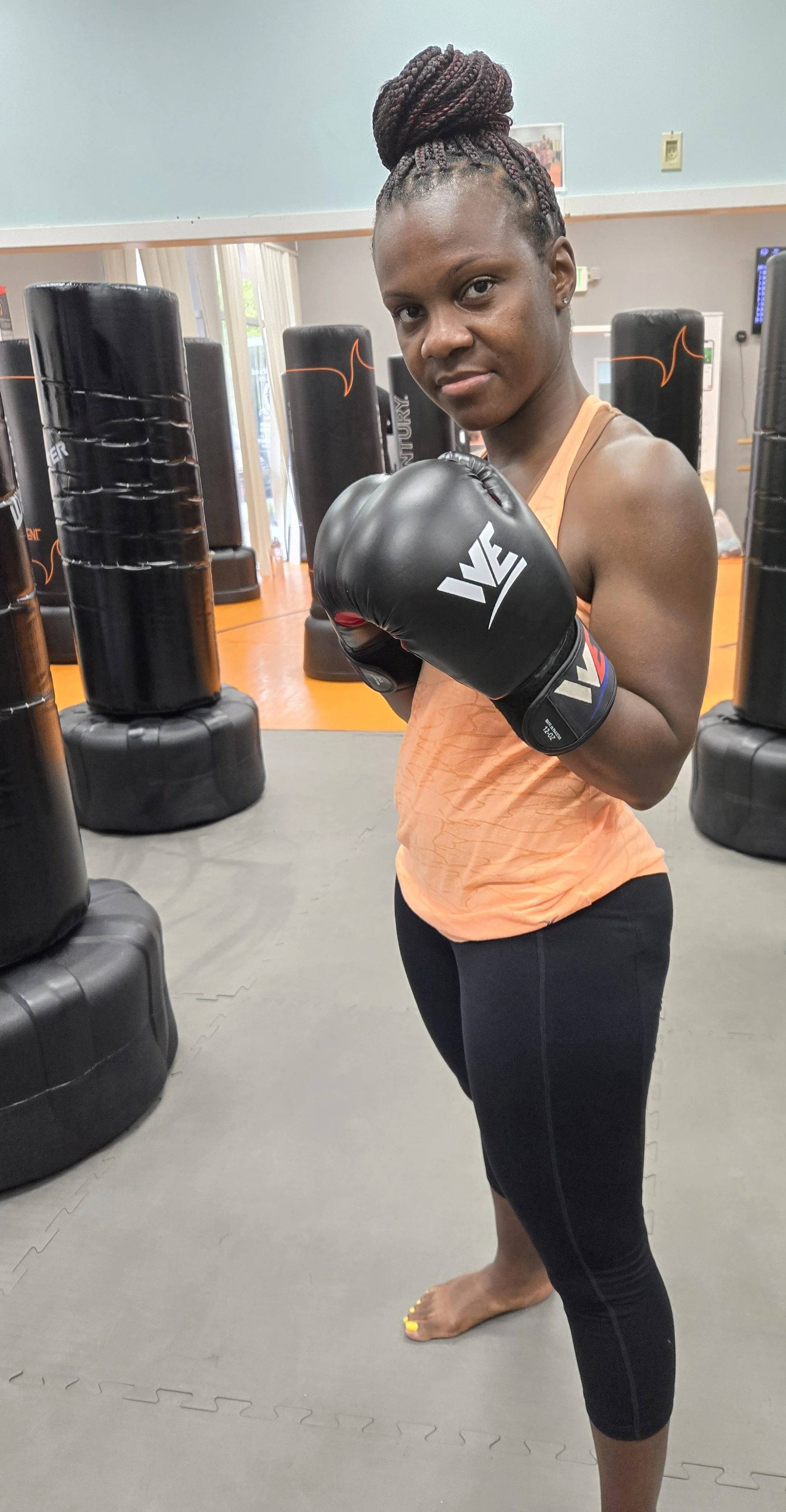 A woman in an orange tank top and black leggings with yellow toenails posing with boxing gloves in a gym with punching bags.