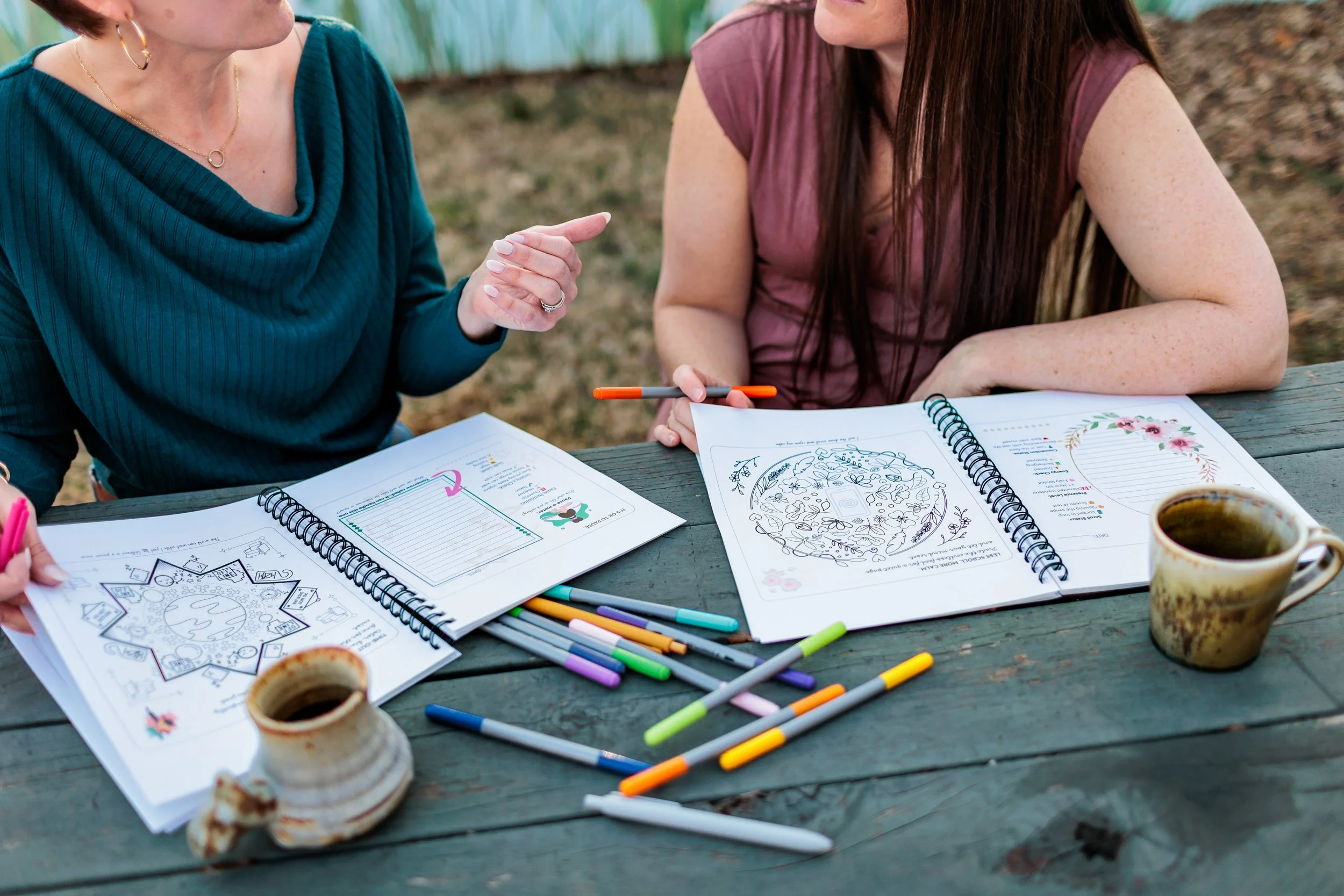 Two women sit at a wooden table outdoors, engaged in a conversation. They have open workbooks filled with drawings and notes, colored markers scattered on the table, and two small cups of coffee. One woman wears a teal top and the other a maroon top.