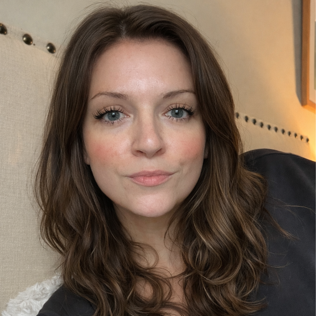 Close-up of a young woman with long, wavy brown hair and blue eyes, smiling softly in a well-lit room with beige walls and a decorative headboard.