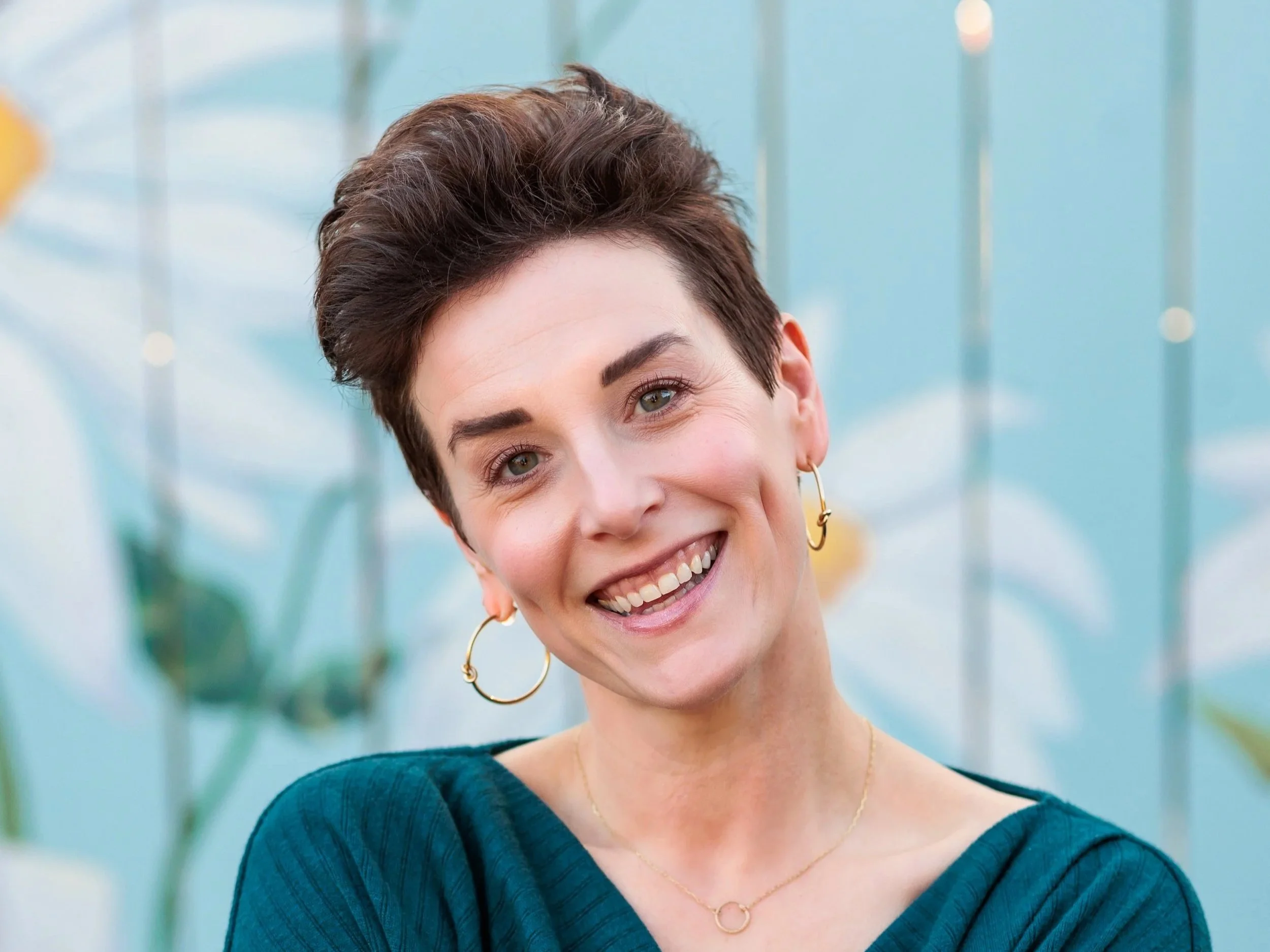 A smiling woman with short dark hair, wearing hoop earrings and a navy blue top, standing in front of a blurred background of glass structures.