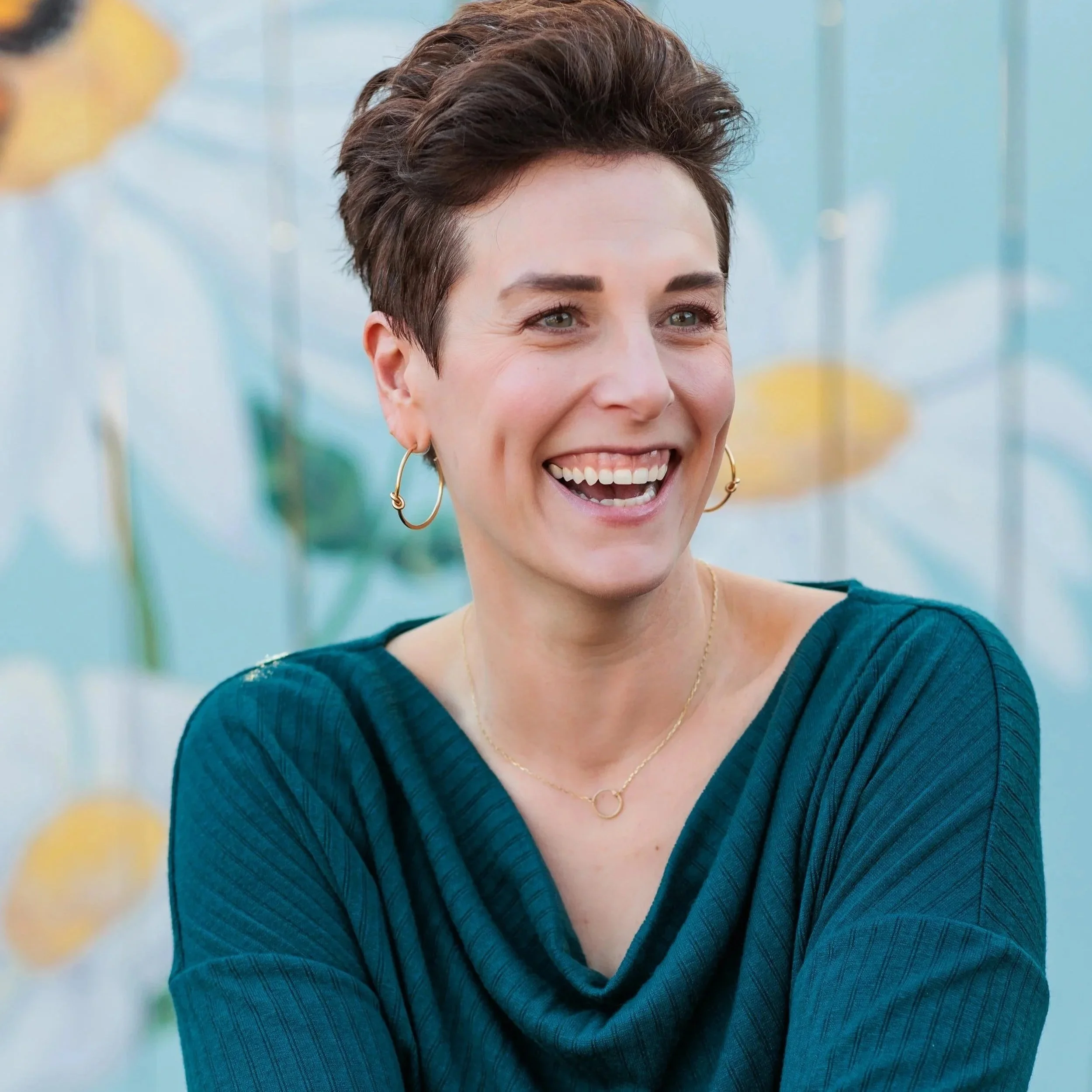A woman smiling with short, dark hair wearing hoop earrings and a teal, ribbed top. The background is softly out of focus with hints of yellow and green.