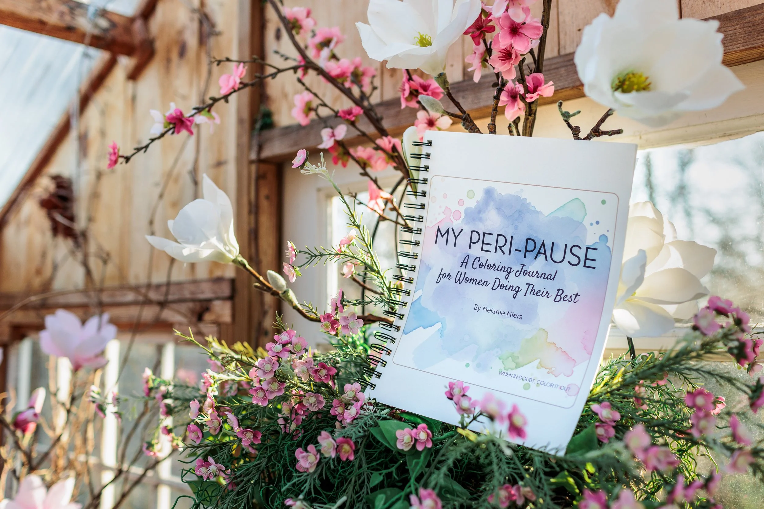 A journal titled "My Peri-Pause" surrounded by pink and white flowers on a rustic wooden surface with sunlight coming through a window.
